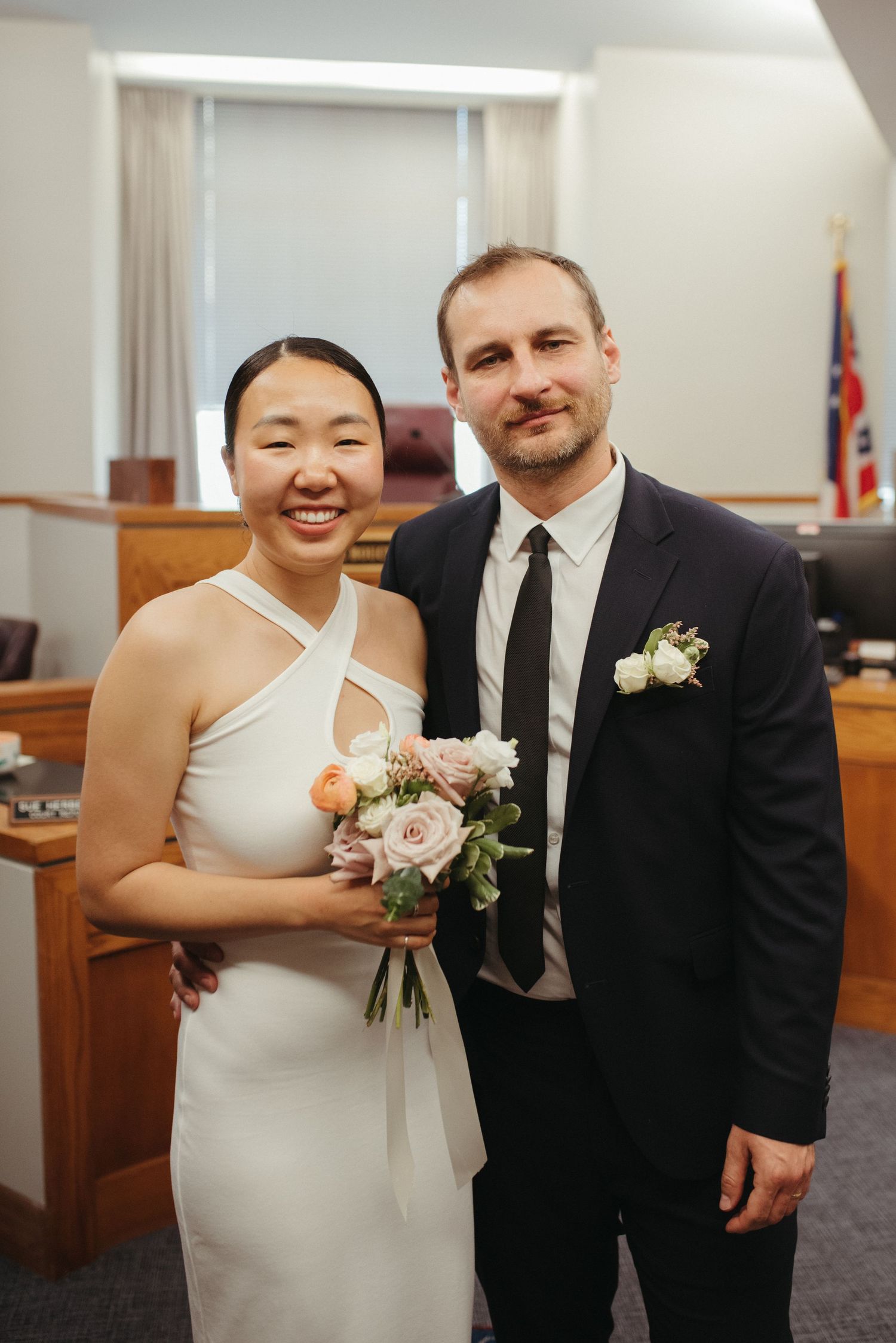 A wedding couple poses in a courthouse, with the bride in a white halter dress holding a pink bouquet.