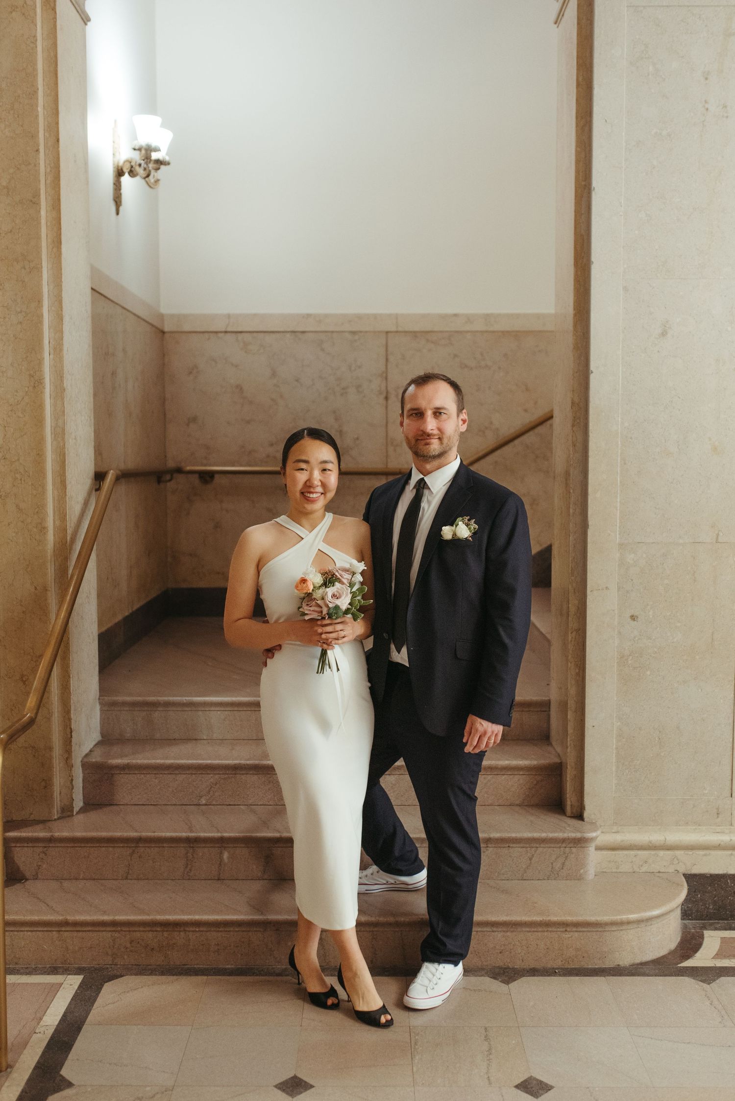 A newlywed couple stands on marble stairs, with the bride in a chic white dress and groom in a dark suit.