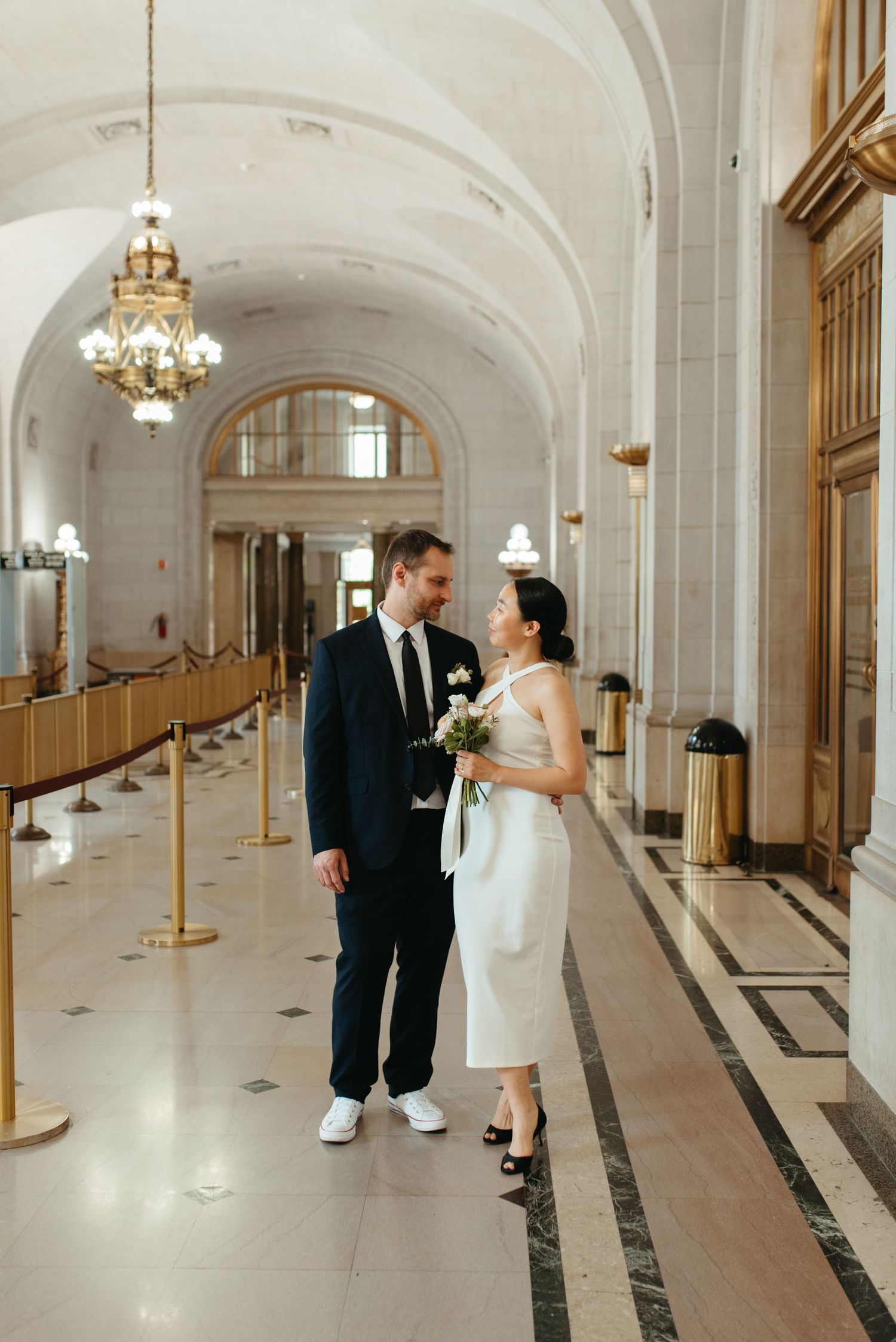 Wedding couple stands in an elegant hallway with arched ceilings and chandeliers.