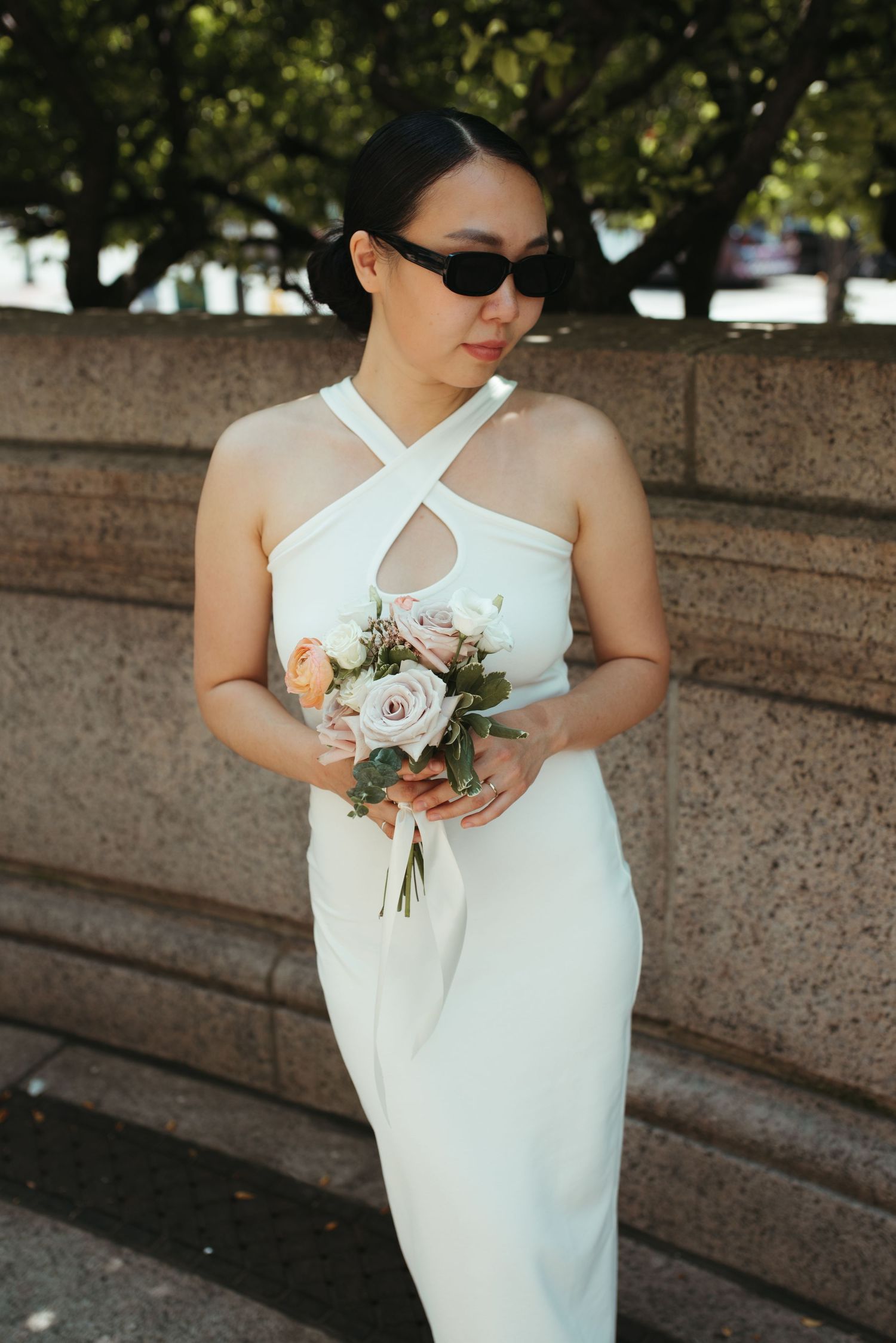 Bride in sunglasses holds a pink rose bouquet against a stone wall backdrop.