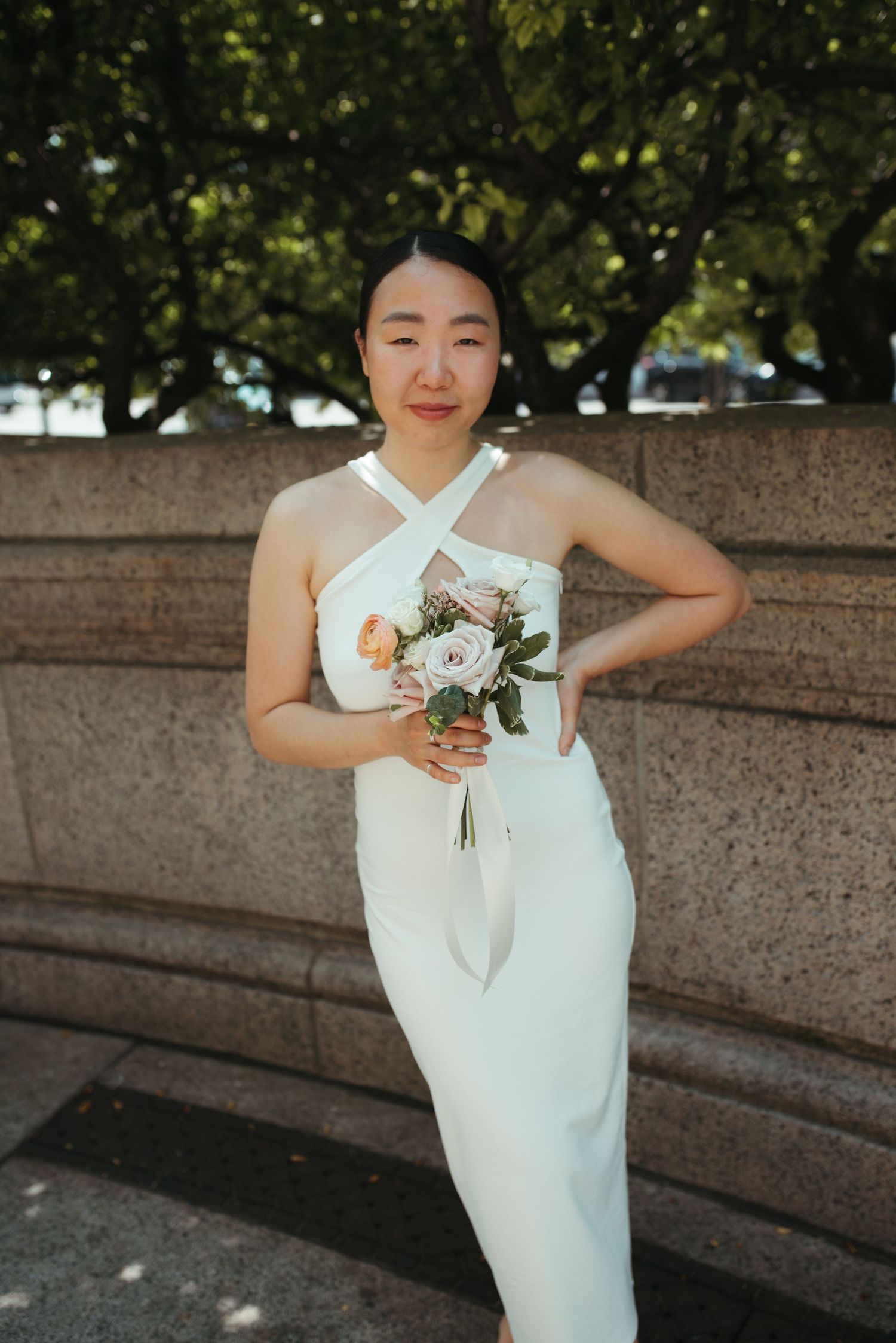 A bride in a sleek white halter dress poses with her bouquet of peach and white roses.
