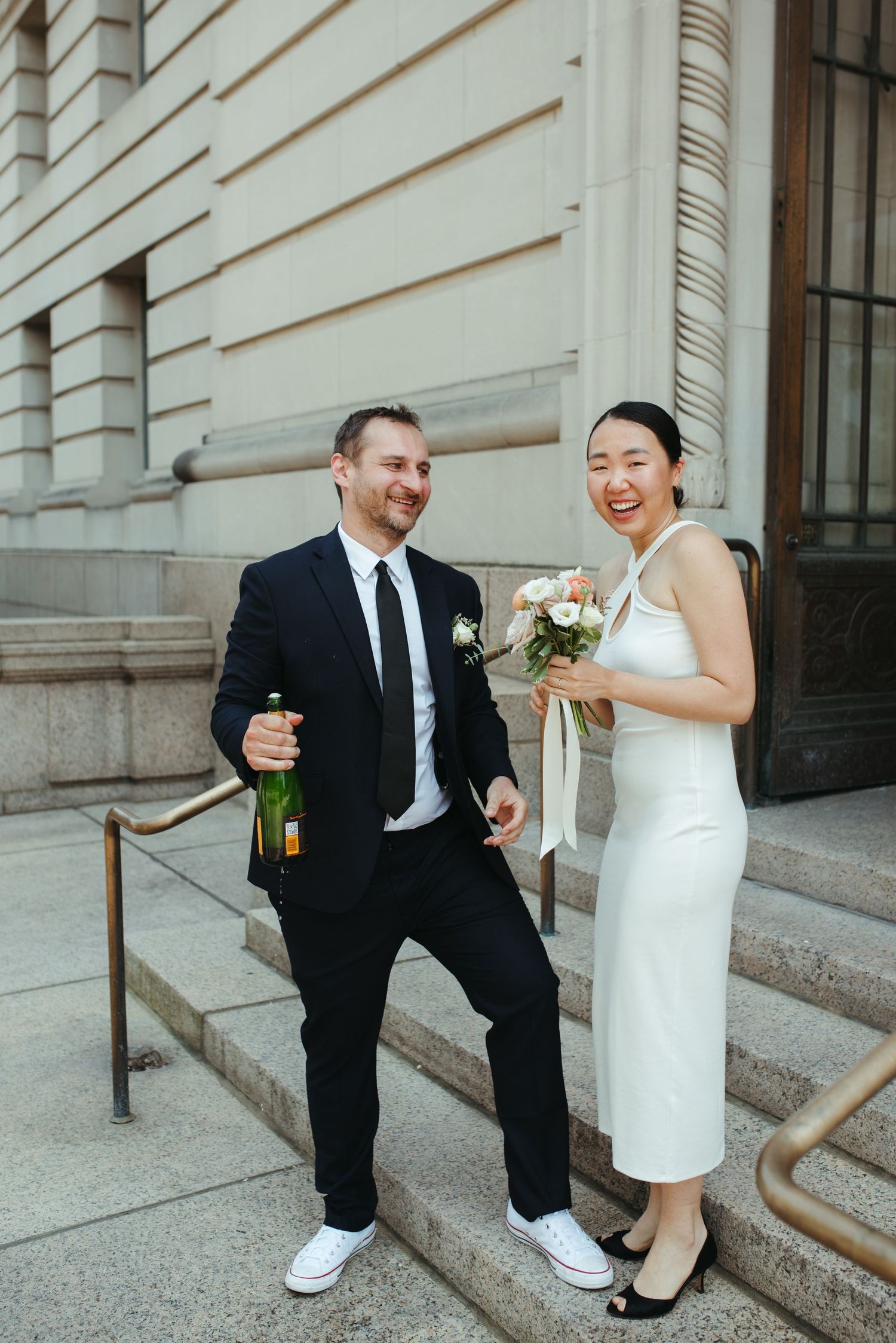 A celebratory moment outside a historic building with the couple holding champagne and flowers.