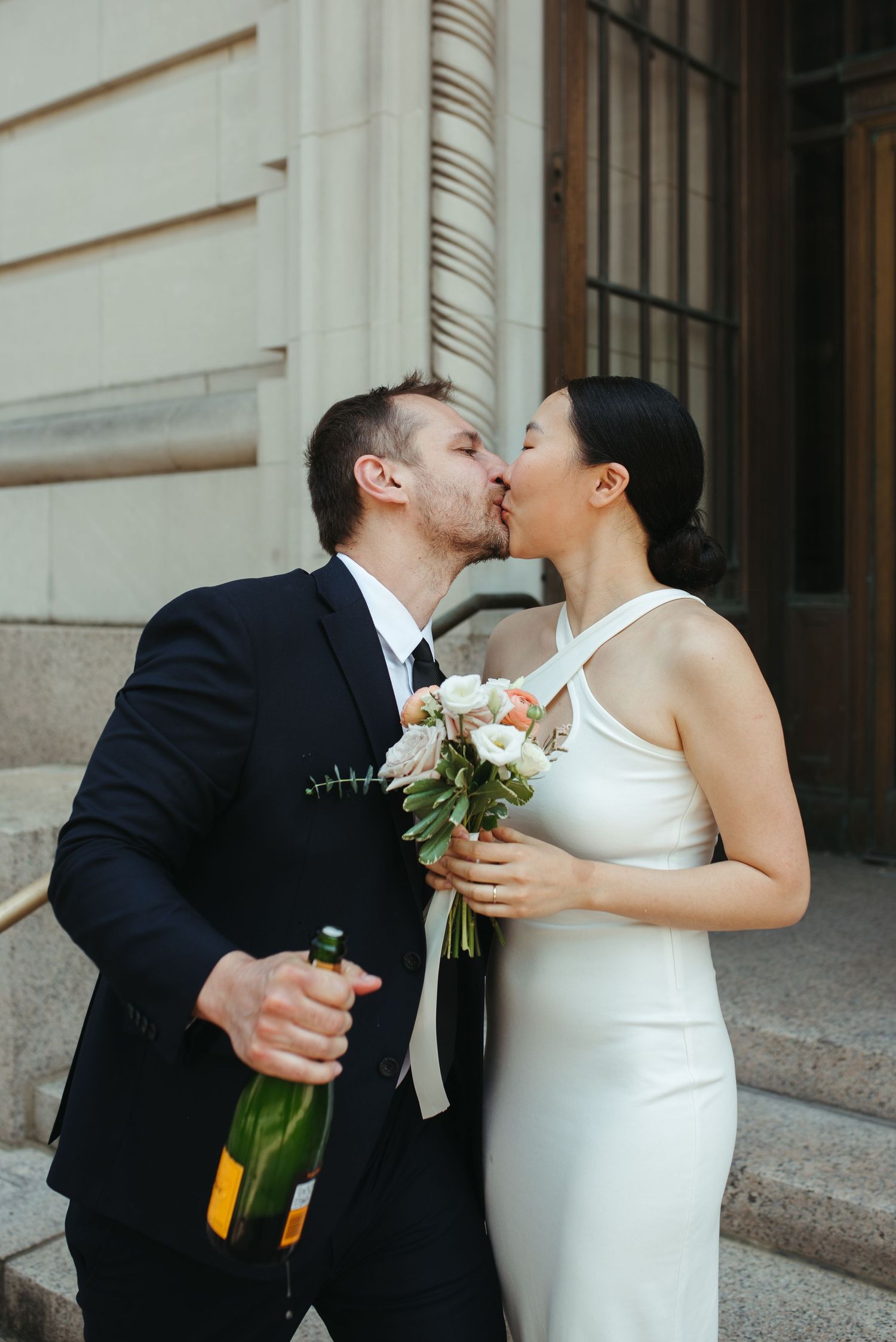 A wedding couple shares a kiss on stone steps while holding champagne and a floral bouquet.