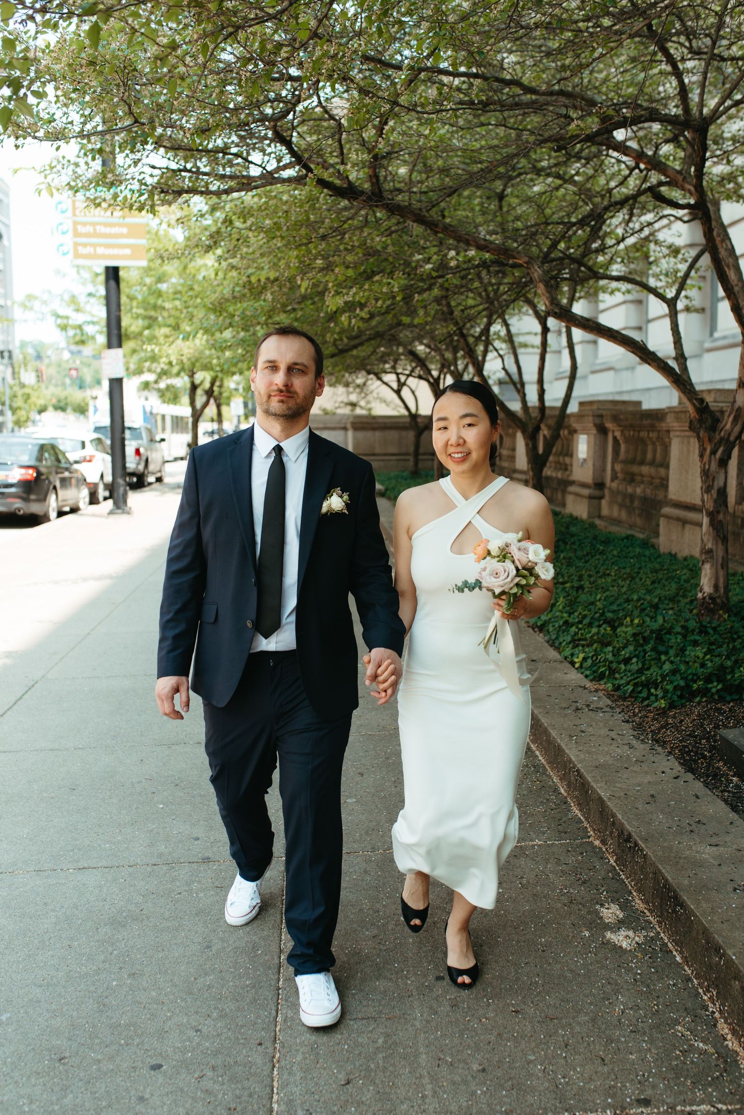 Newlyweds stroll together on a sunny sidewalk lined with green trees.