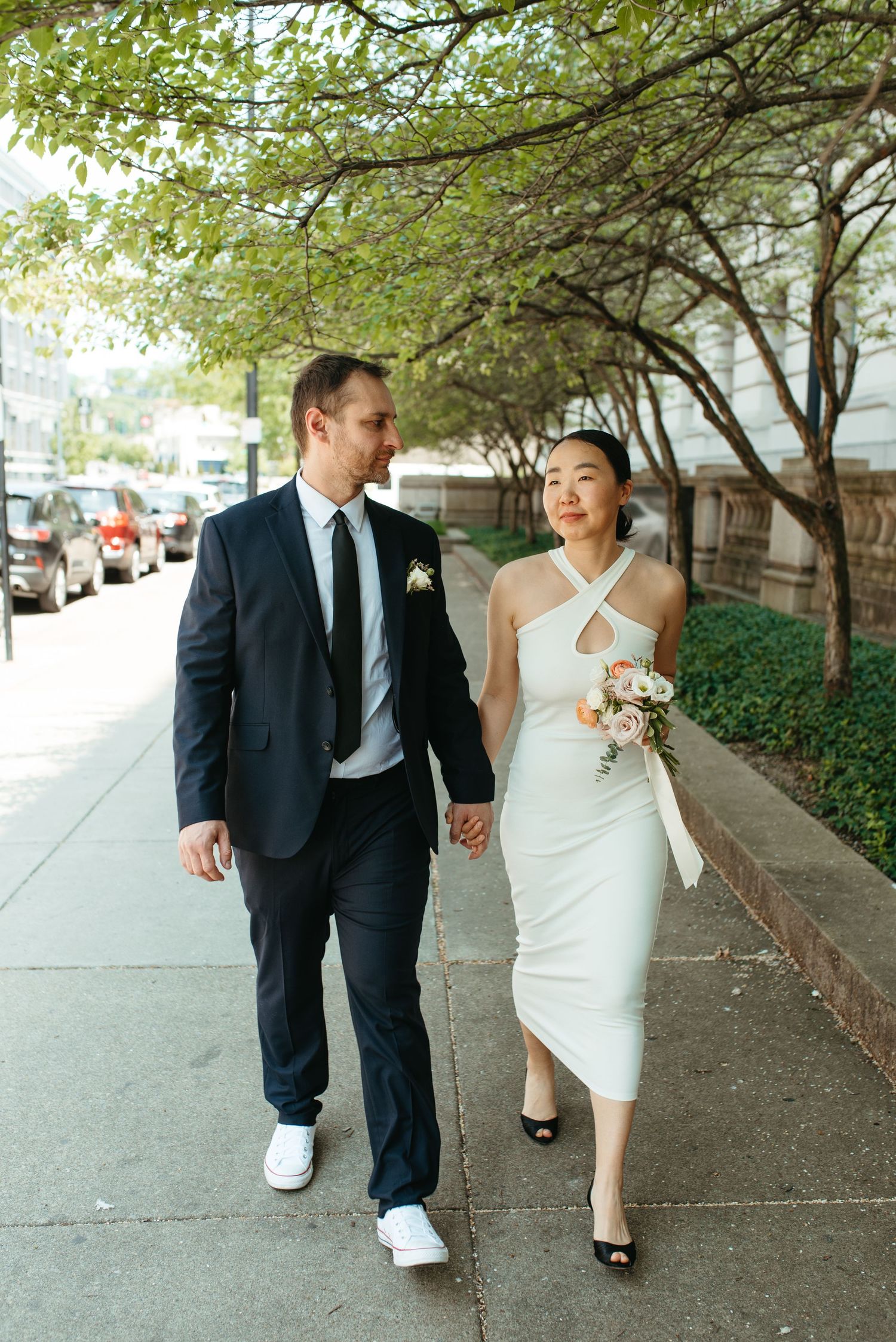 A happy couple walks hand in hand under tree-lined streets after their wedding ceremony.