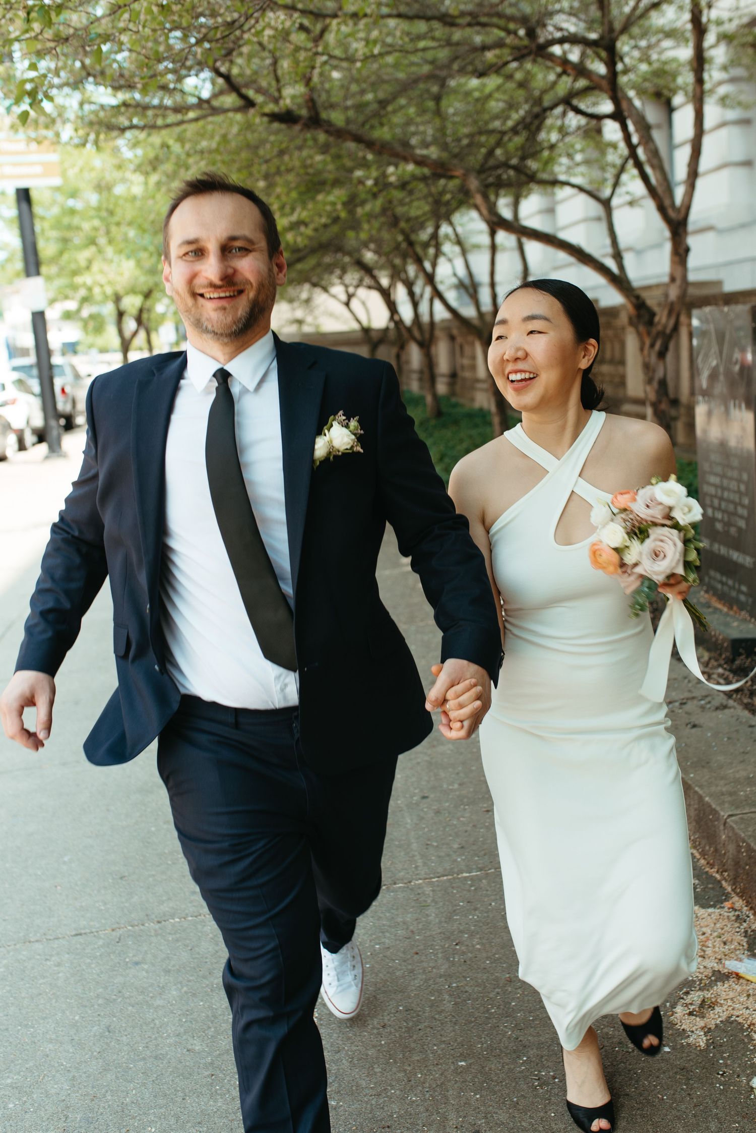 A joyful couple runs hand in hand down a tree-lined urban sidewalk.