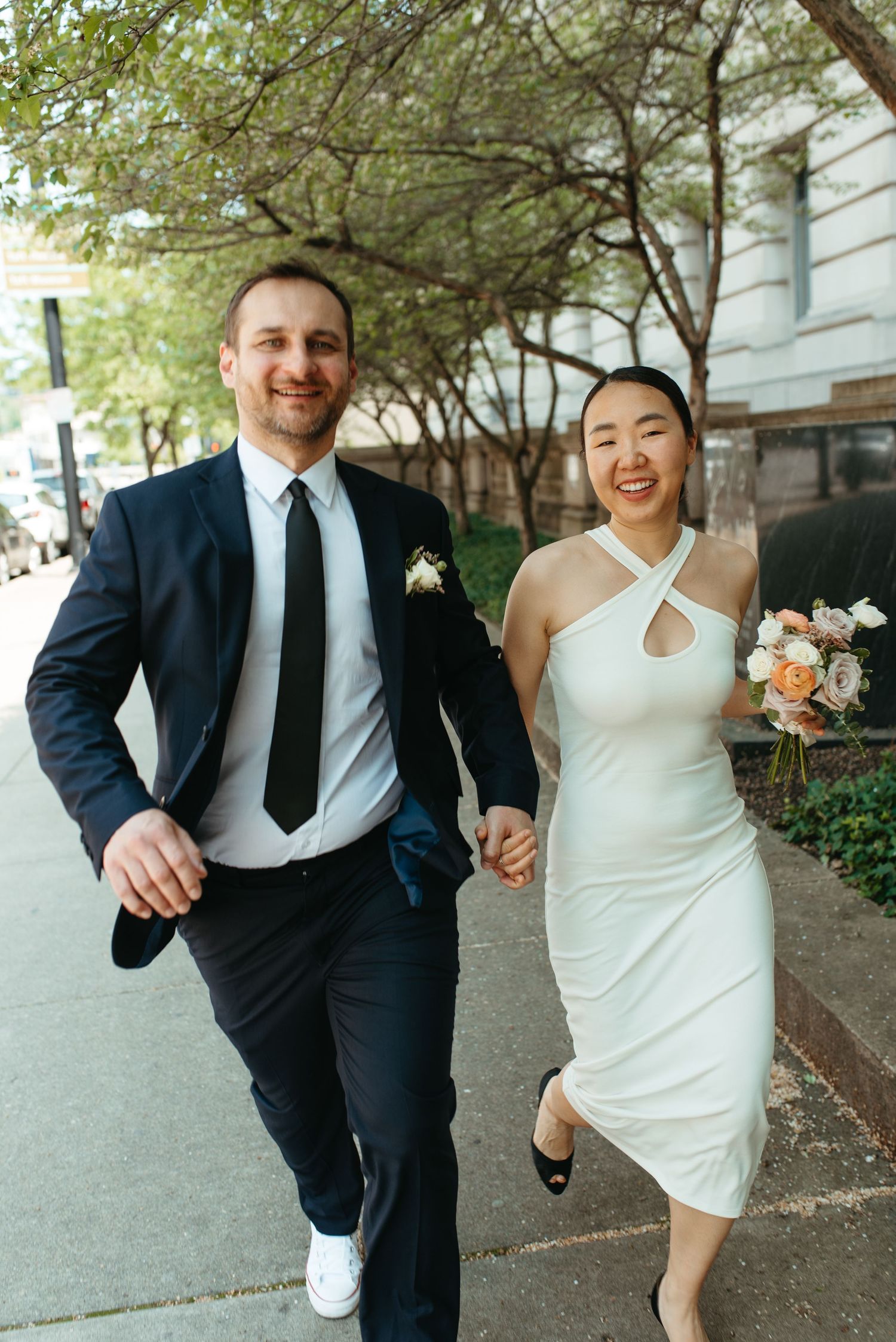 A happy couple laughs while running down a city sidewalk after their wedding ceremony.