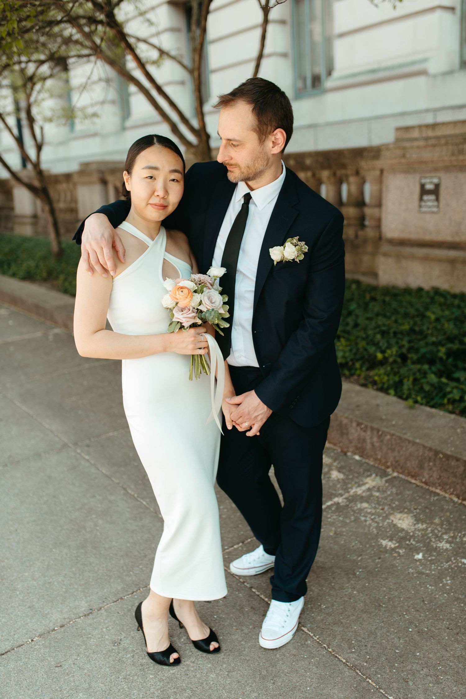 A wedding couple poses together on a city sidewalk, the bride holding a delicate bouquet.