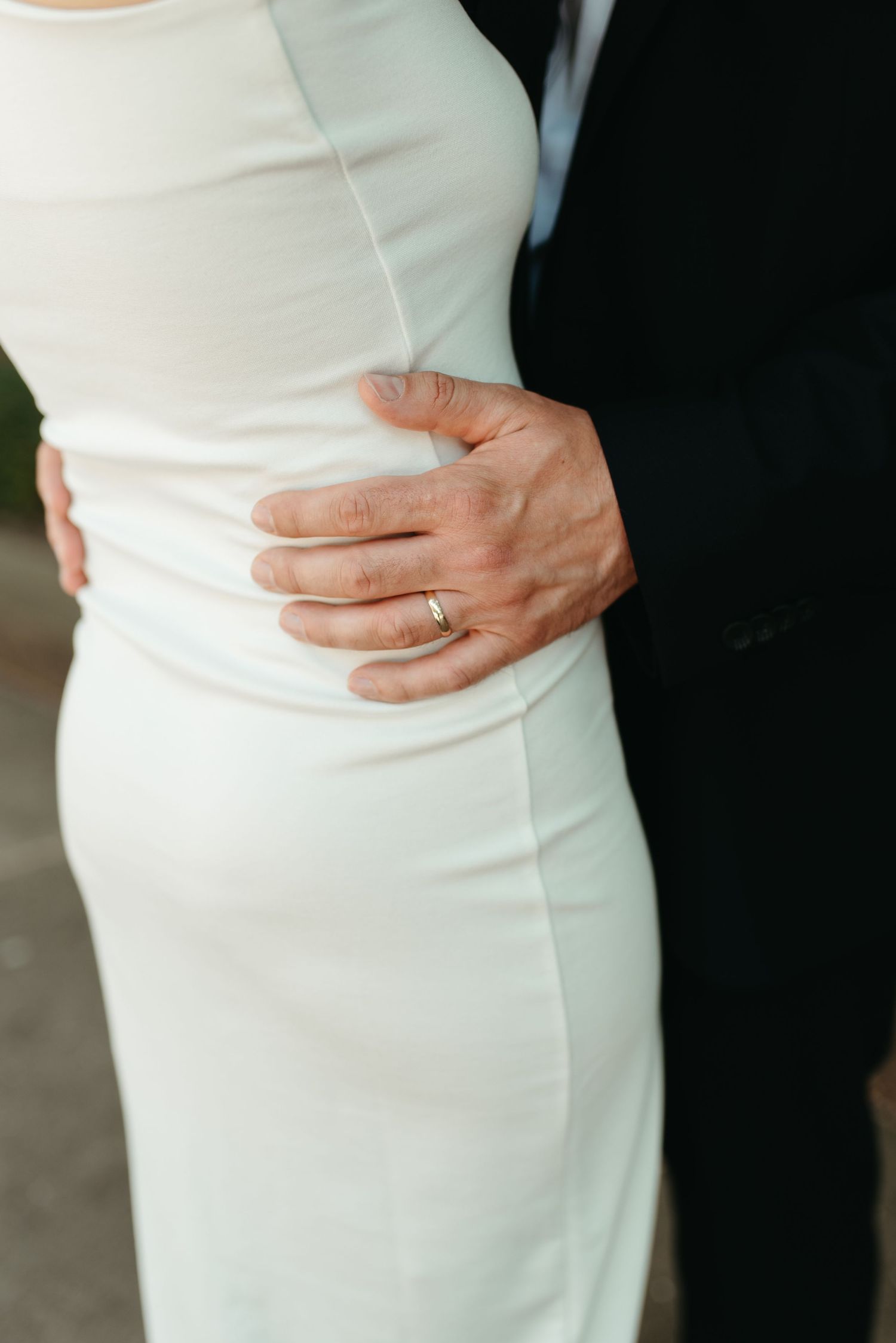 Close-up of a groom's hand with wedding ring resting on bride's white dress.