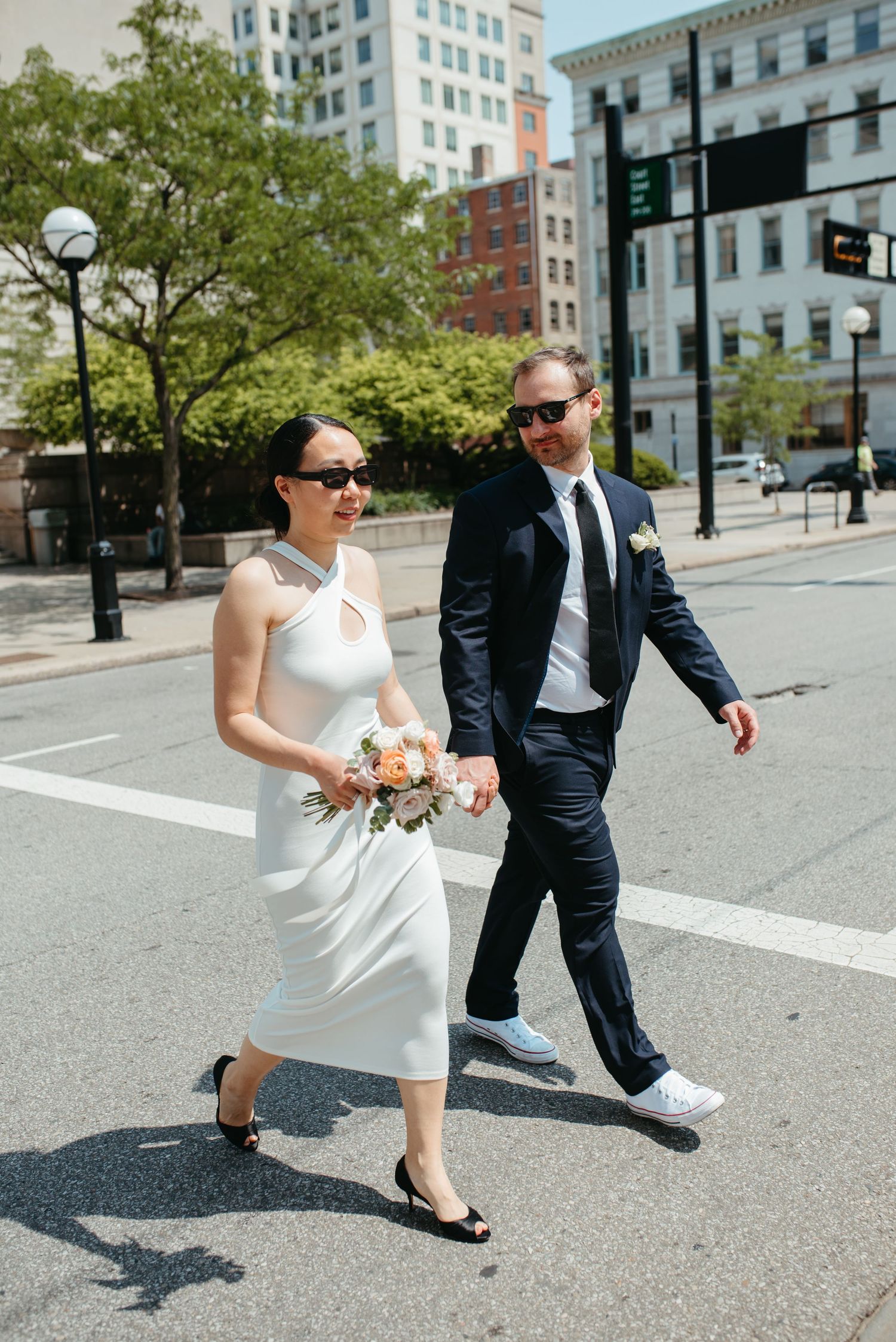 A stylish couple in wedding attire strolls through a sunny city street wearing sunglasses.