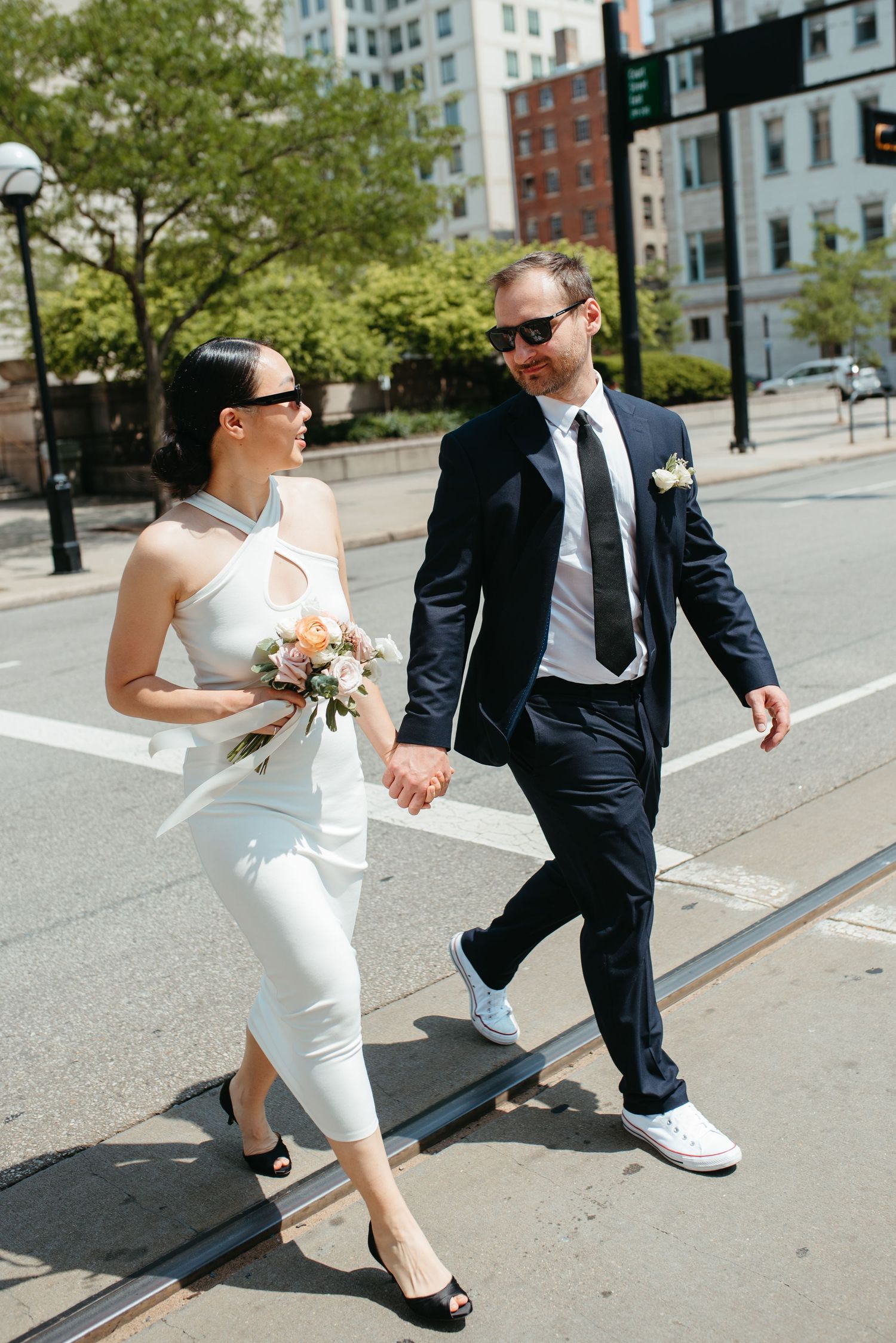 A newlywed couple walks hand in hand down a sunny urban street.