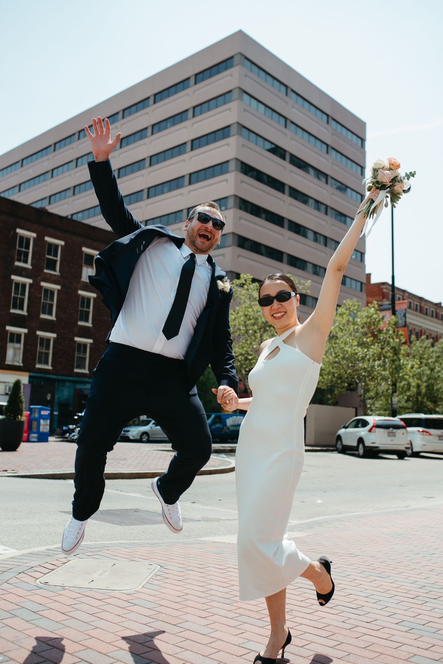 A joyful wedding couple jumps with excitement in front of a city building.