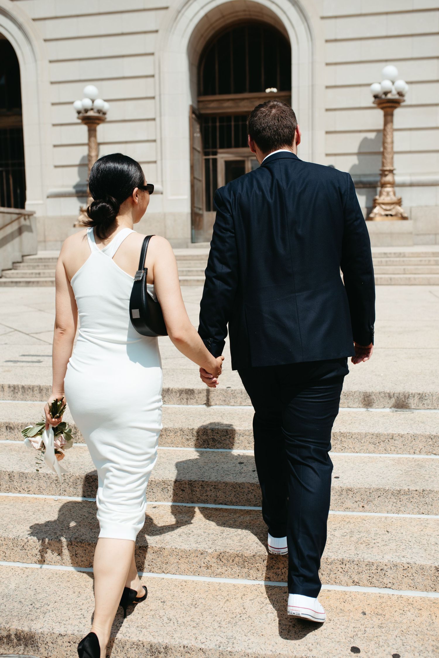 Couple in formal attire ascends stone courthouse steps while holding hands.