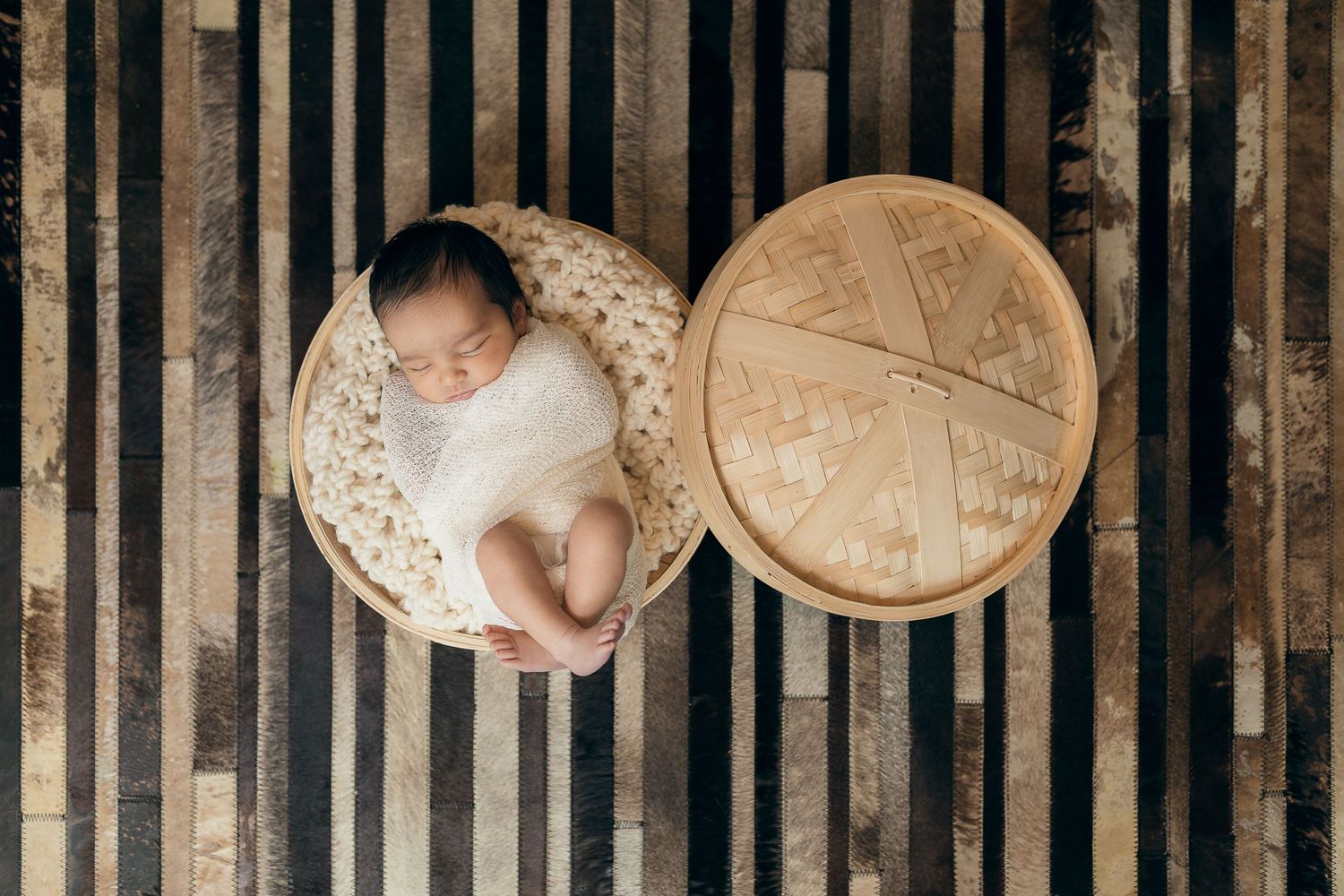 Overhead view of sleeping baby in white swaddle in a bamboo dim sum steamer on striped surface.