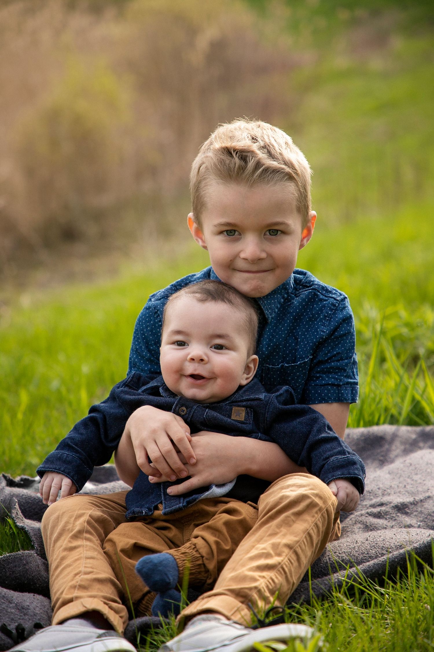 a boy with his baby brother in his lap sitting on a blanket in a grassy field