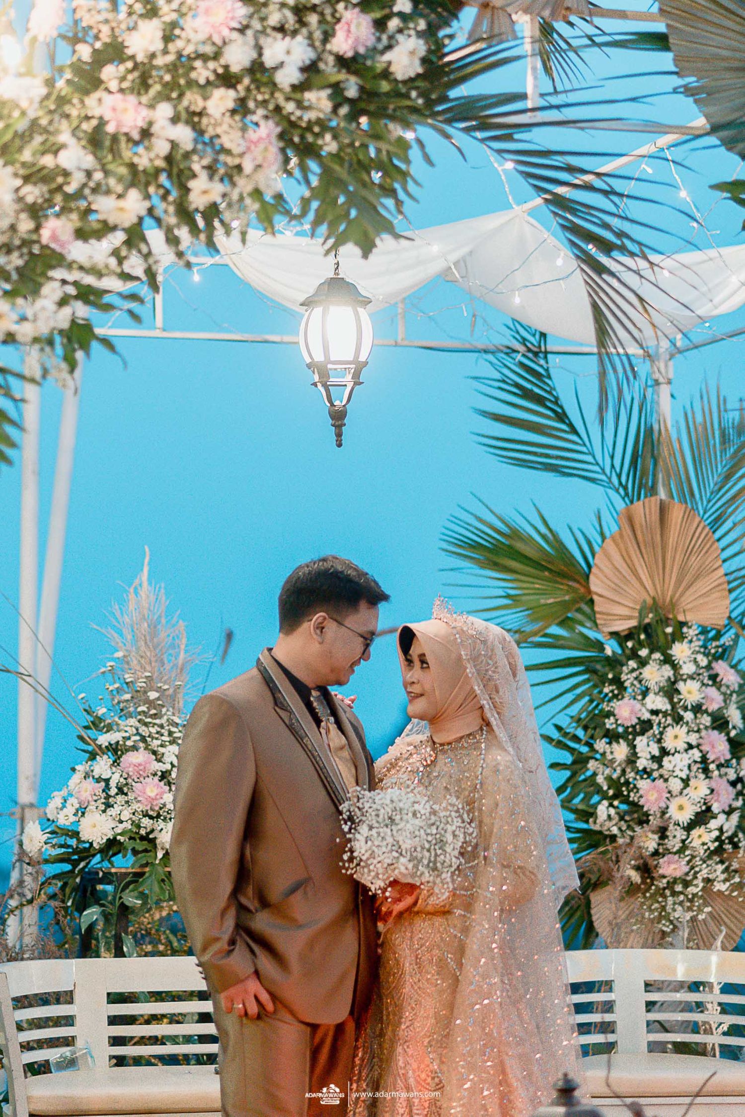 Romantic wedding portrait under palm leaves and hanging lanterns against blue sky.