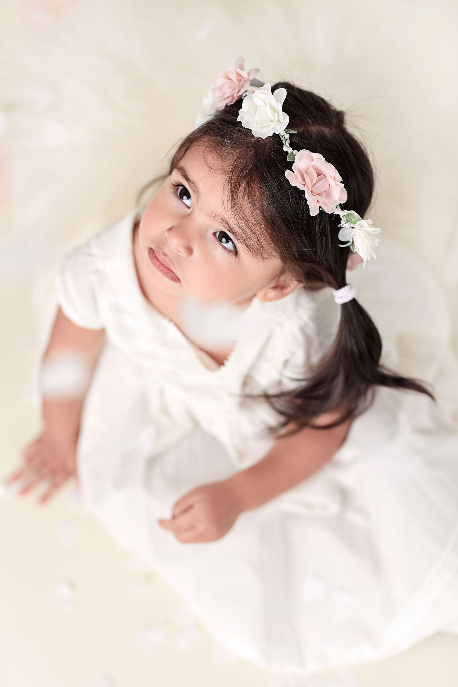 A small child in a white dress wears a delicate floral crown with pink and white flowers.