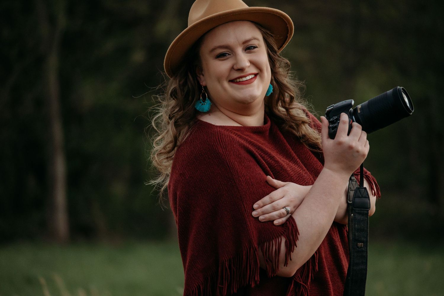 A photographer in a burgundy poncho and brown hat smiling while holding a camera outdoors.