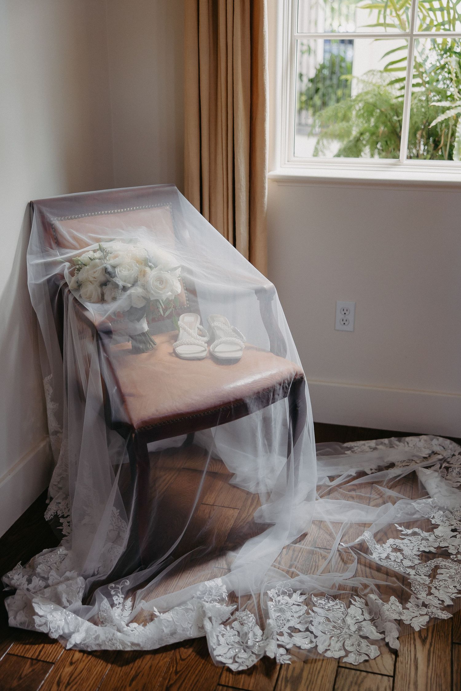 White wedding veil draped over a chair near a window with bridal accessories and lace details on hardwood floor.