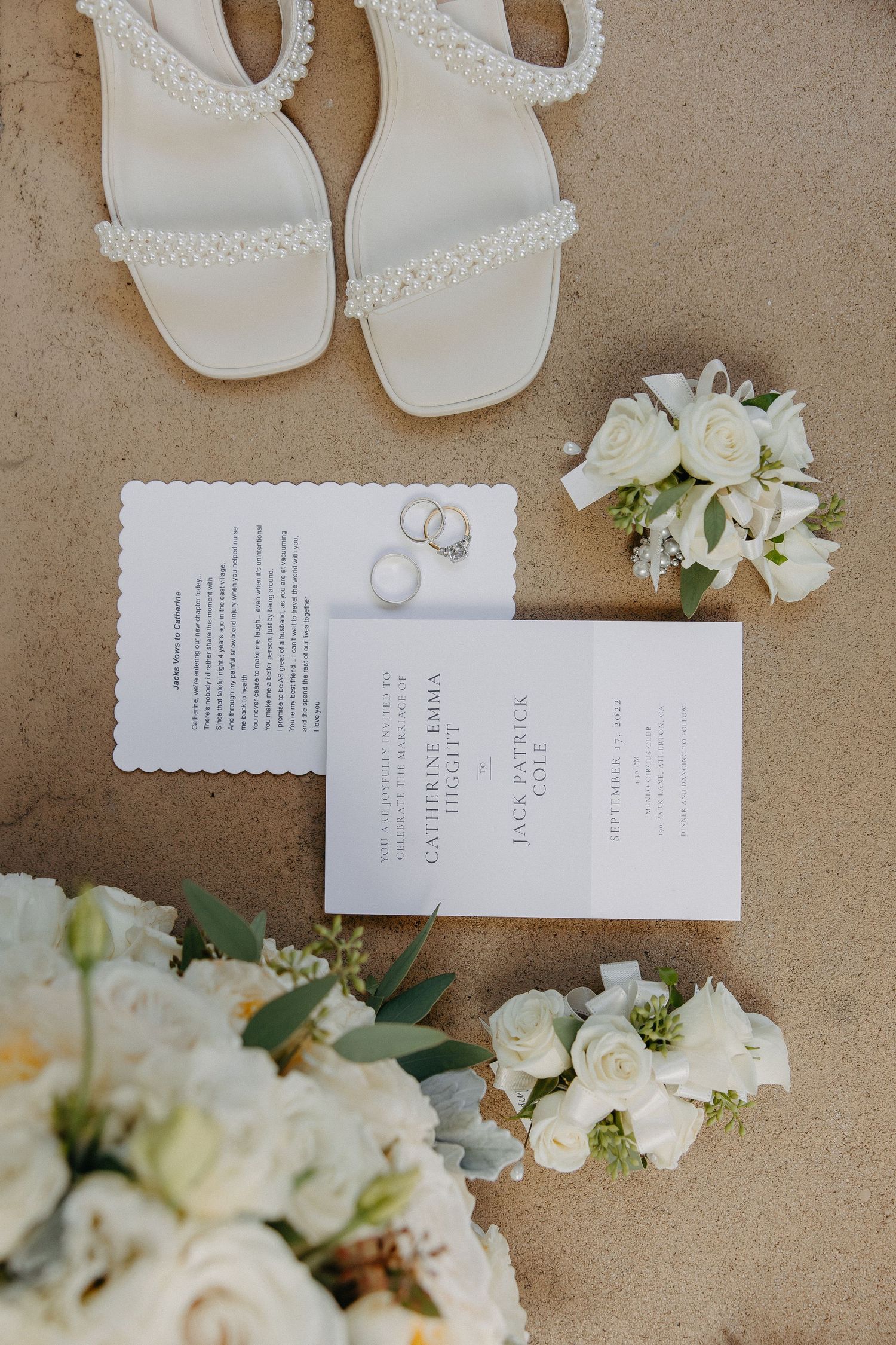 Wedding details flatlay with white sandals, invitation cards, rings and white roses on beige background.