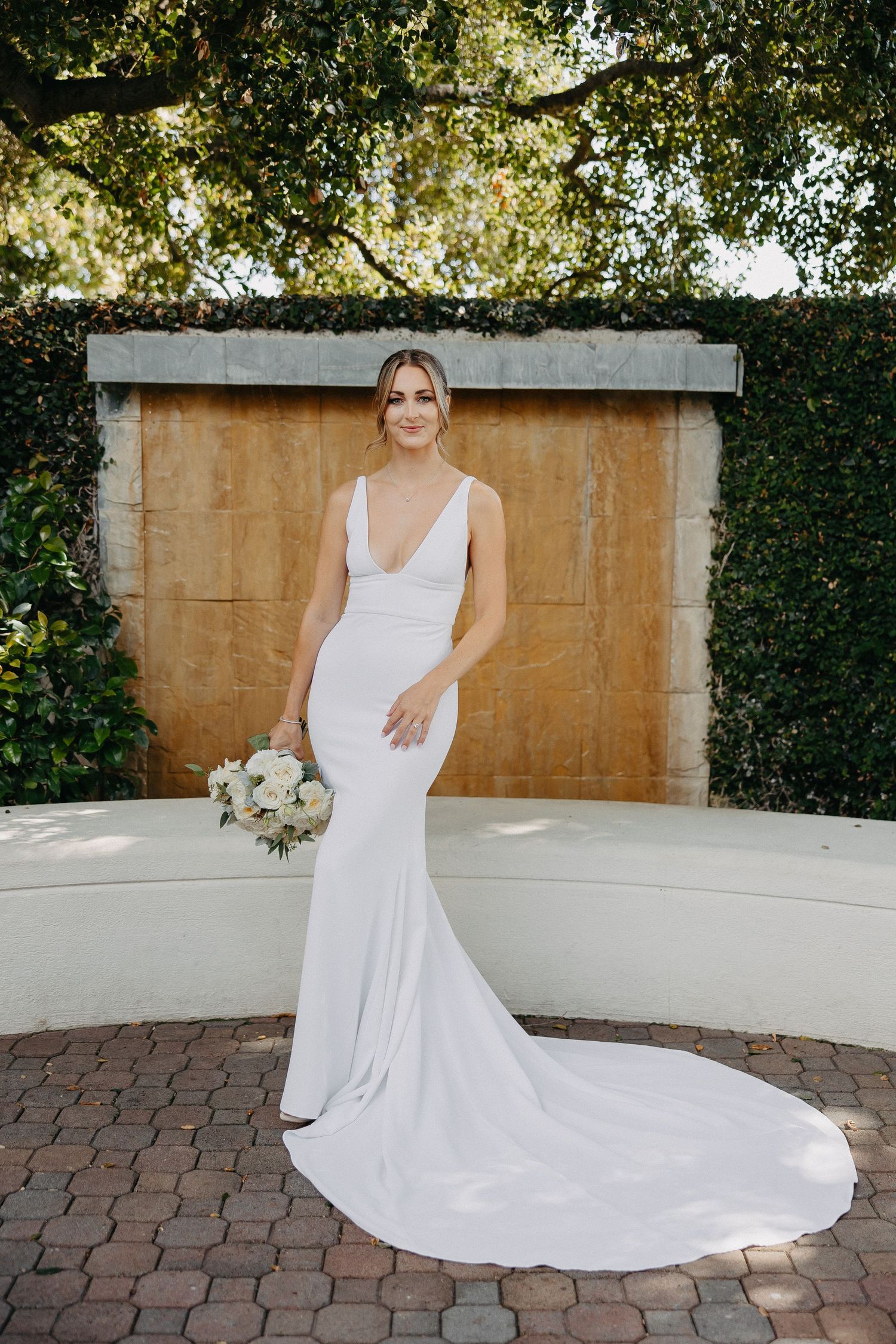 Classic white wedding gown with train posed against stone wall and greenery backdrop.