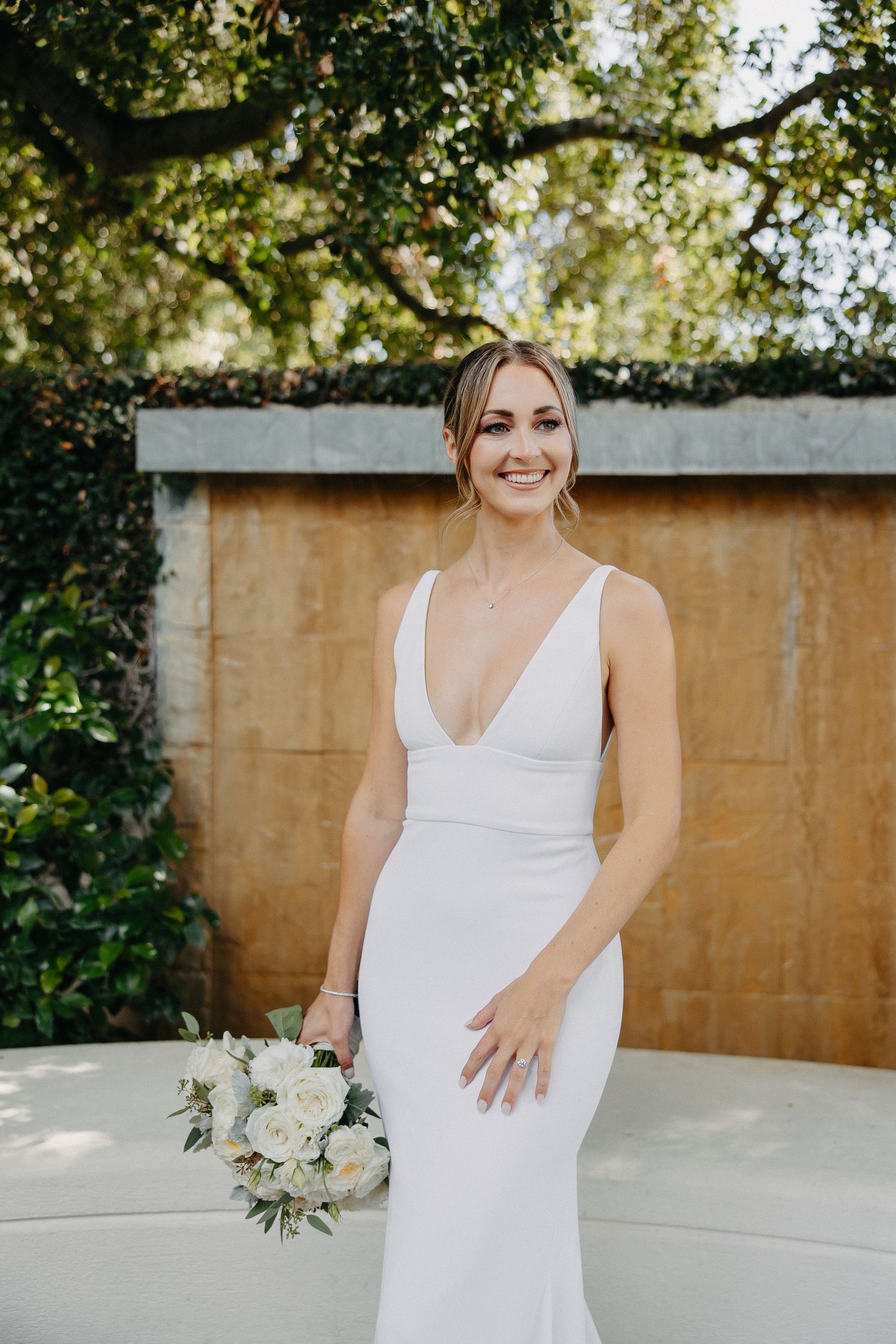 Elegant bridal portrait in fitted white dress with bouquet against garden wall.
