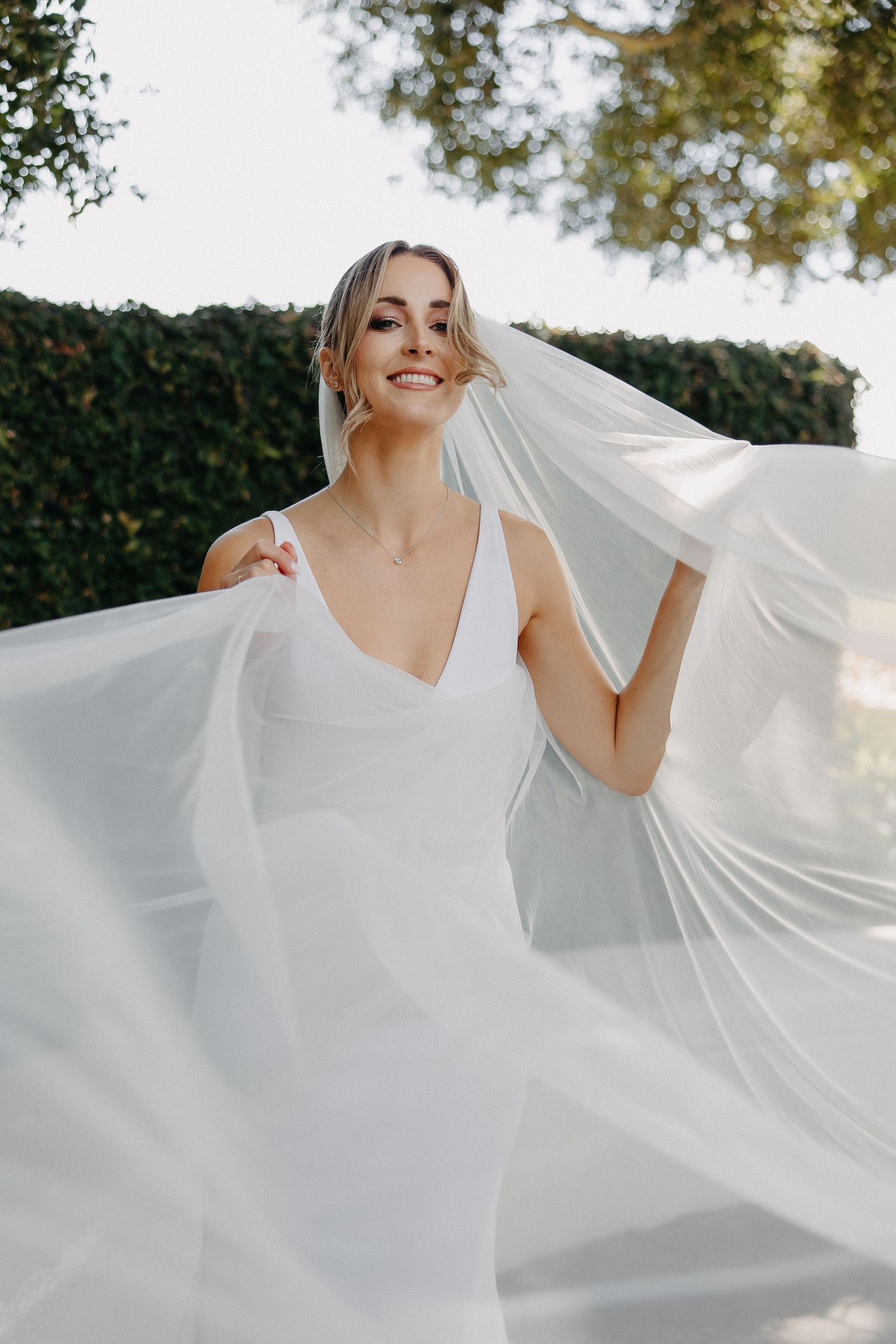 Joyful bridal portrait with floating veil against garden greenery.