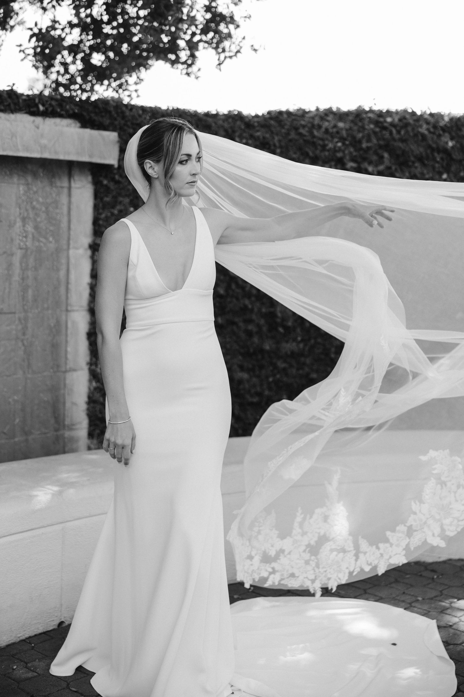 Elegant bride in fitted white gown with flowing veil poses against garden wall in black and white photo.