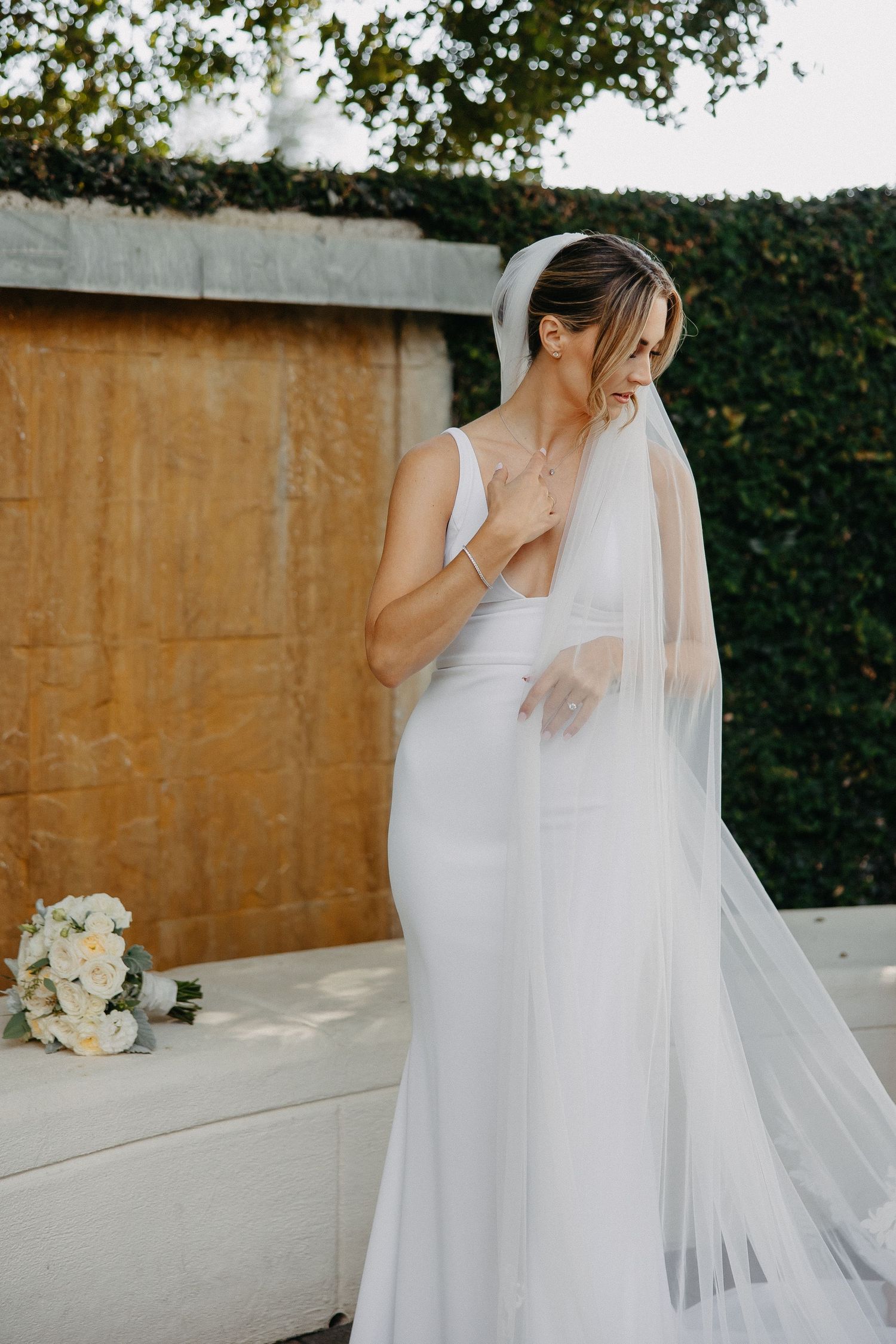 Bride in sleek white dress touches neckline while standing outdoors with flowing veil and bouquet.