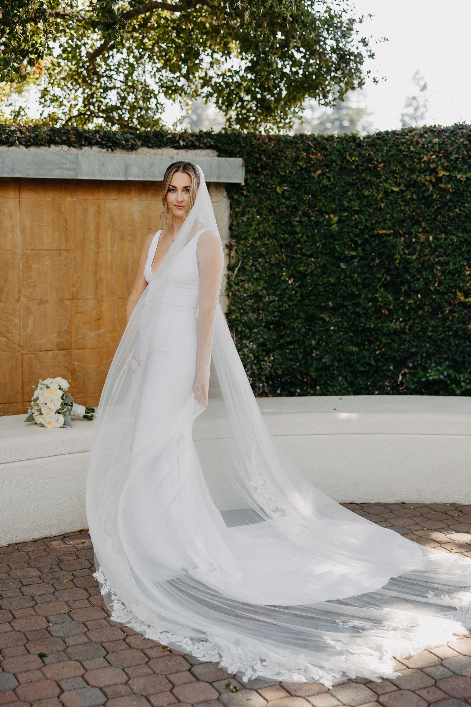 Bride in minimalist white gown with long trailing veil poses by ivy-covered wall.