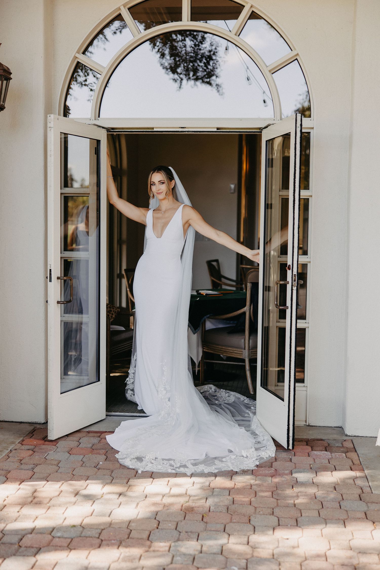 Bride stands in elegant arched doorway showing full length of fitted gown and veil.