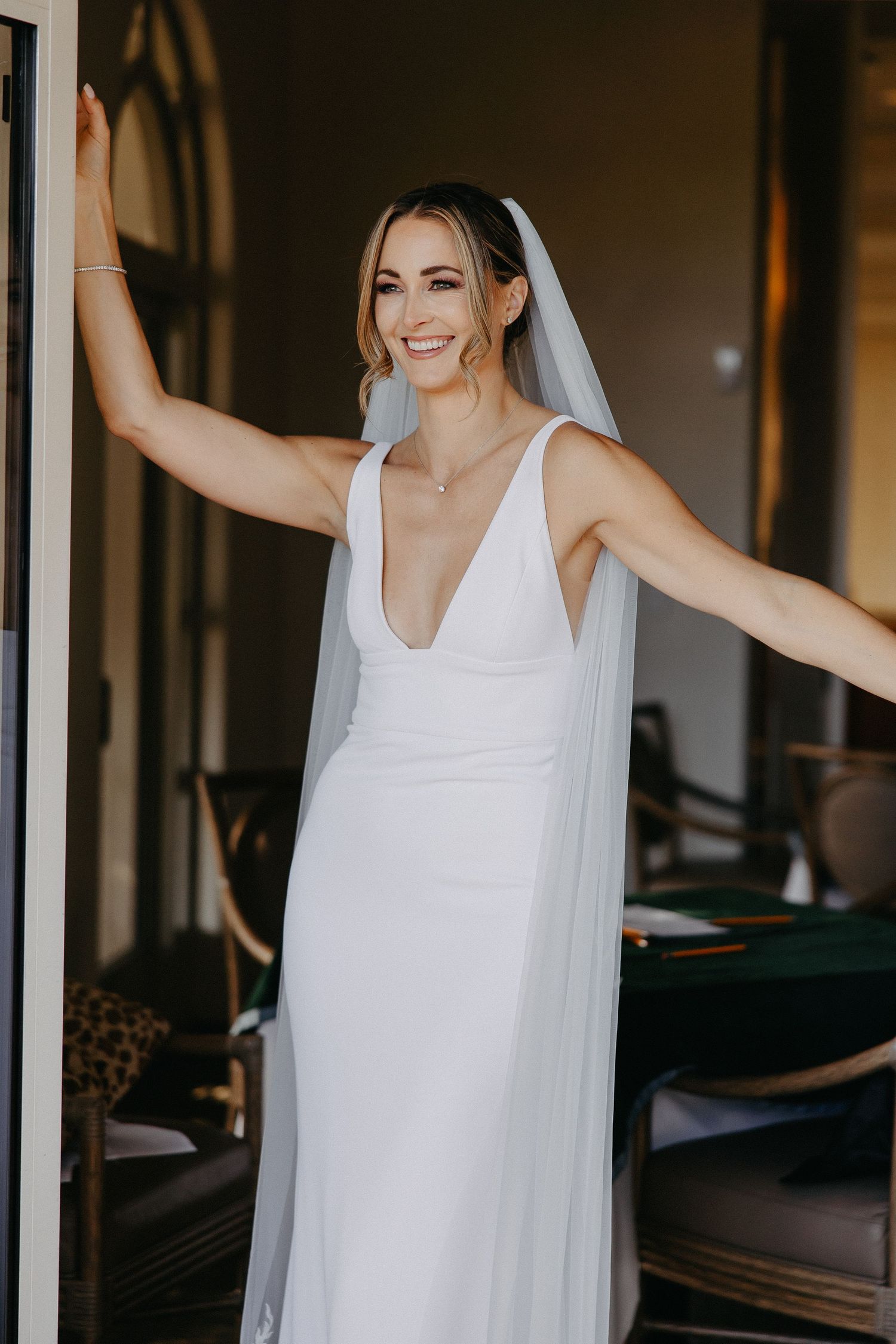 Elegant bride in flowing white v-neck wedding dress smiles joyfully with arms outstretched in doorway.