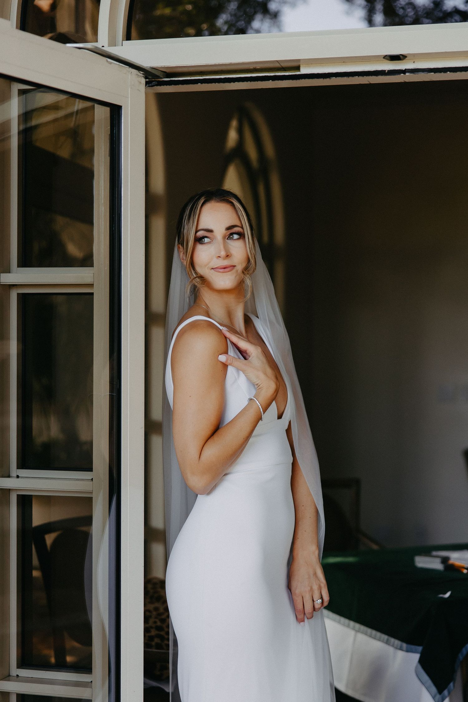 Candid bridal portrait showing sleek white wedding gown and delicate veil in doorway.