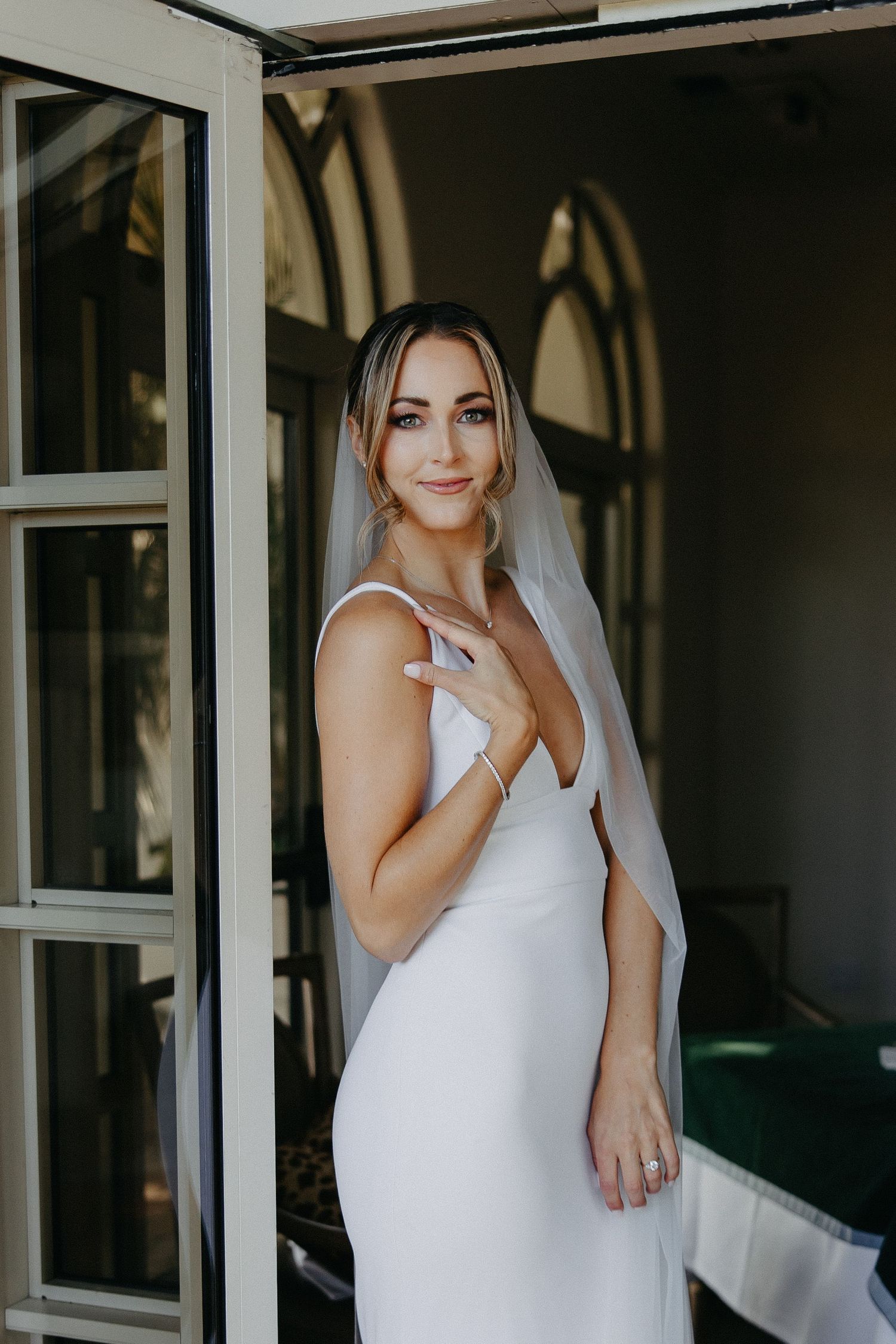 Elegant bride in minimalist v-neck wedding dress poses gracefully near French doors.