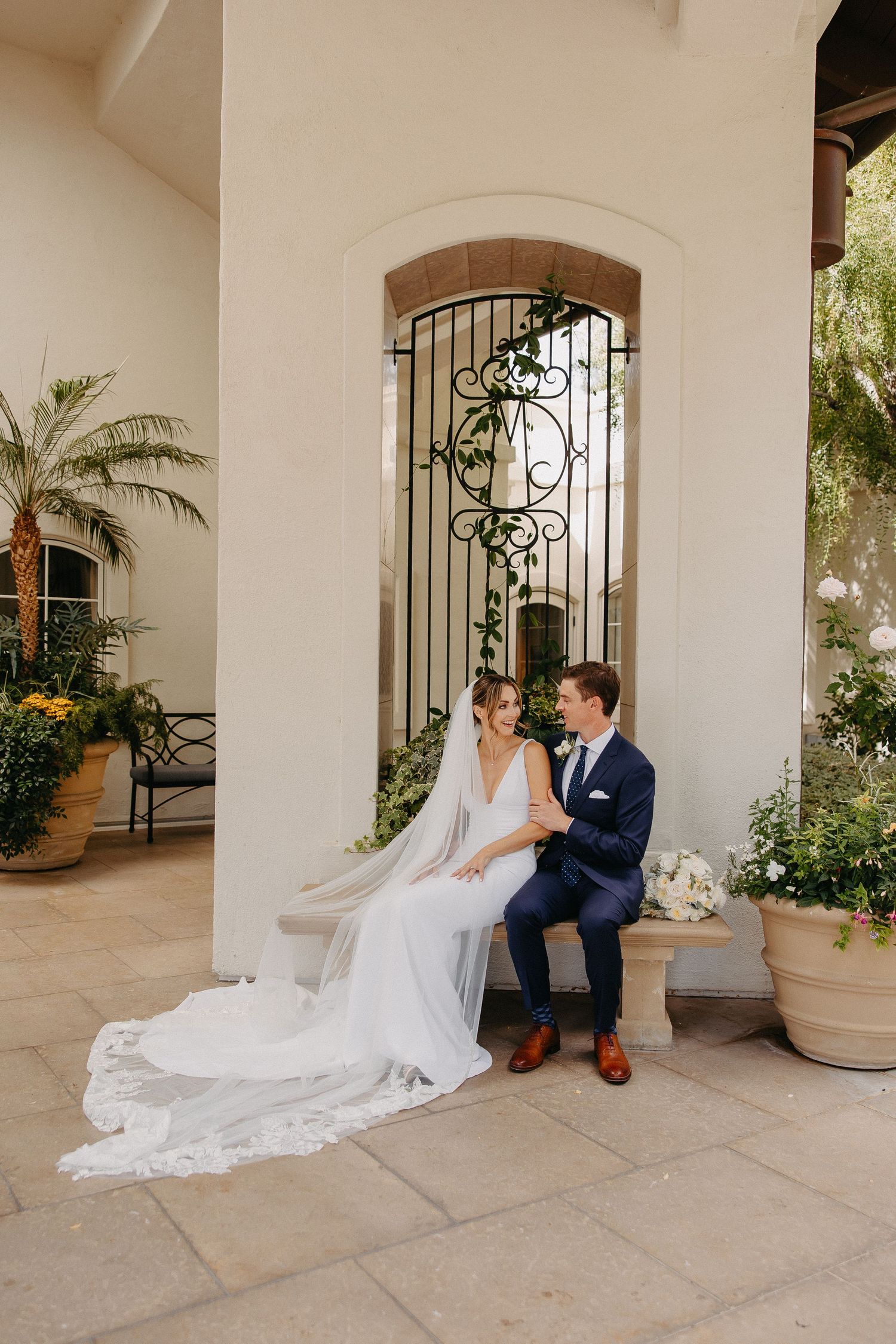 Newlyweds share intimate moment on ornate stone patio with decorative iron gate backdrop.