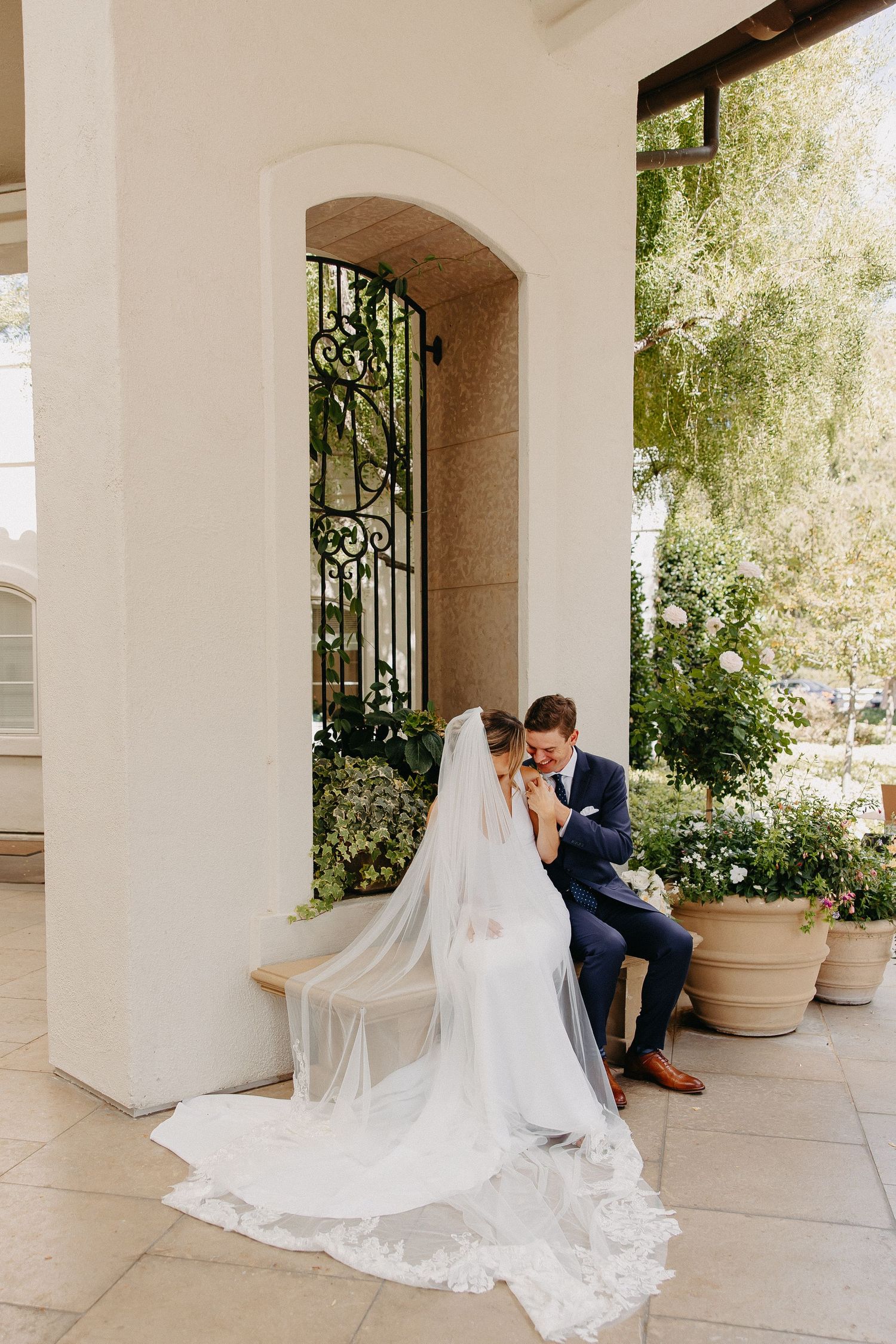 Bride in flowing white gown sits with groom on stone steps by decorative iron gate.