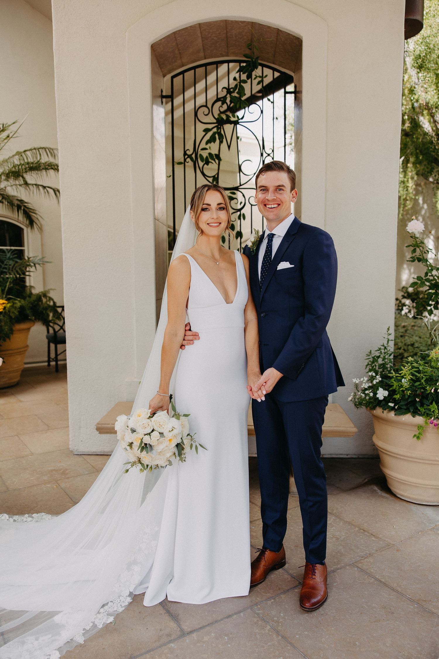 Elegant wedding couple poses in front of Mediterranean-style archway with flowing bridal veil.