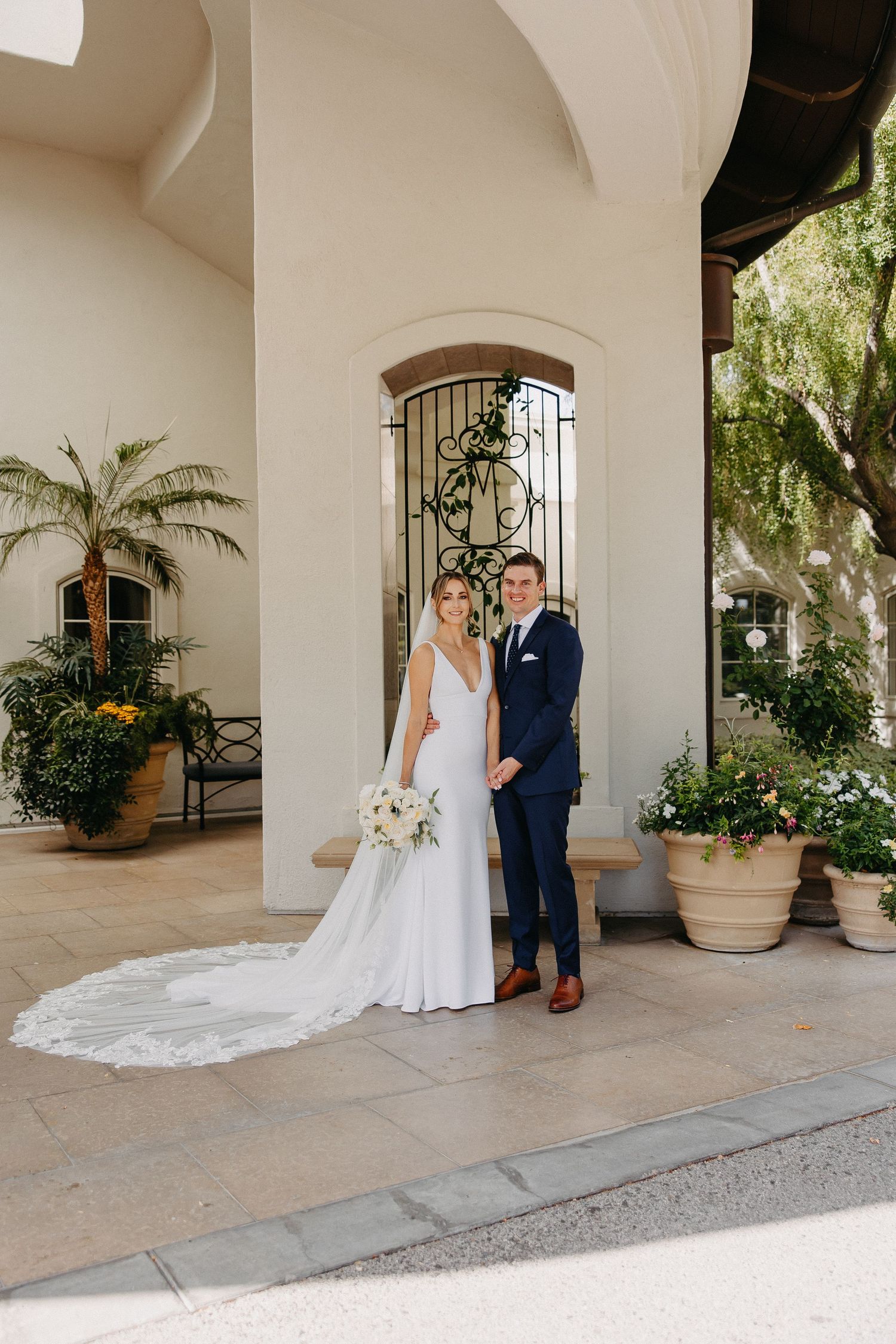 Bride in sleek white gown and groom in navy suit pose by Mediterranean architecture.