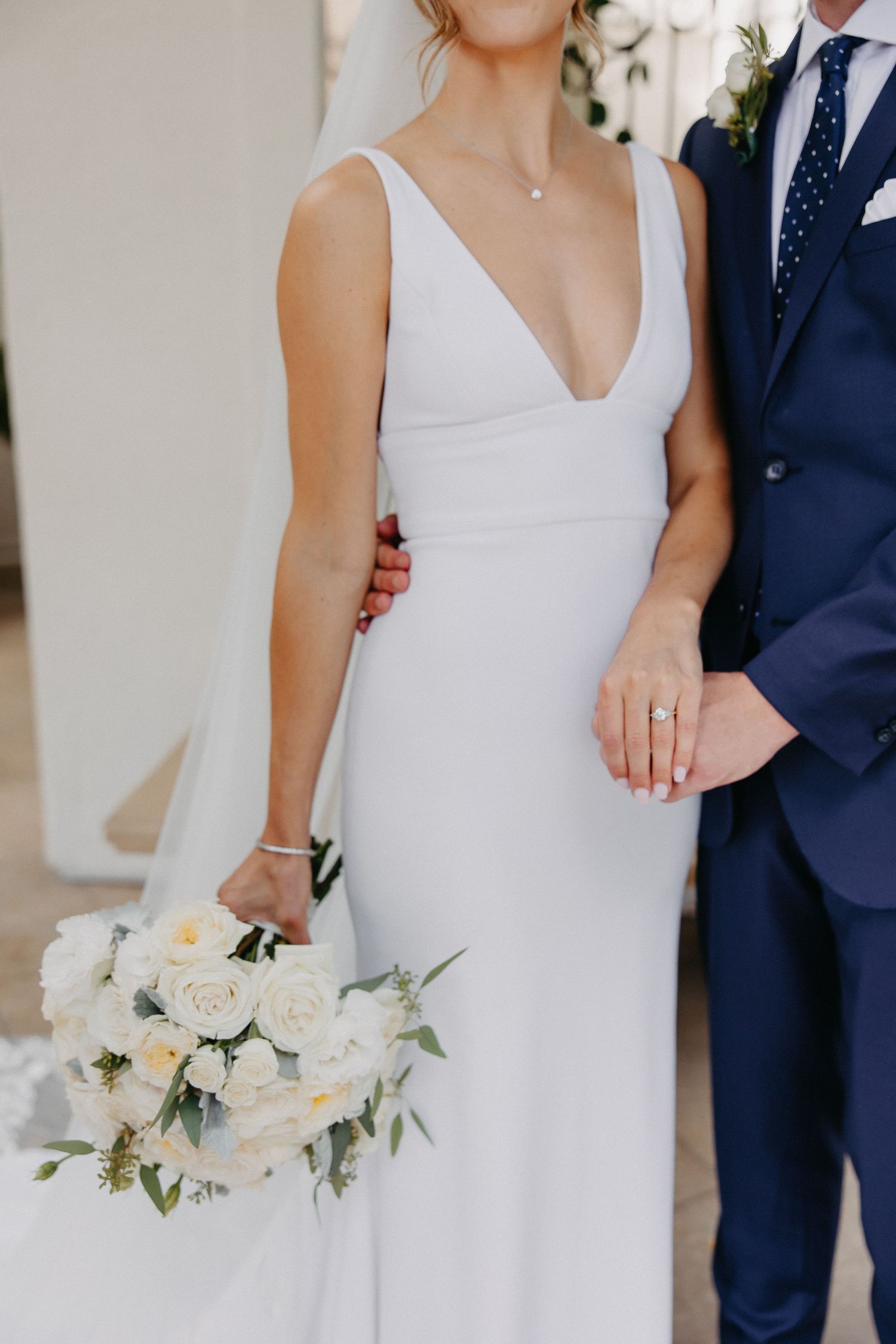 Close-up of bride's white v-neck dress and white rose bouquet next to groom's navy suit.