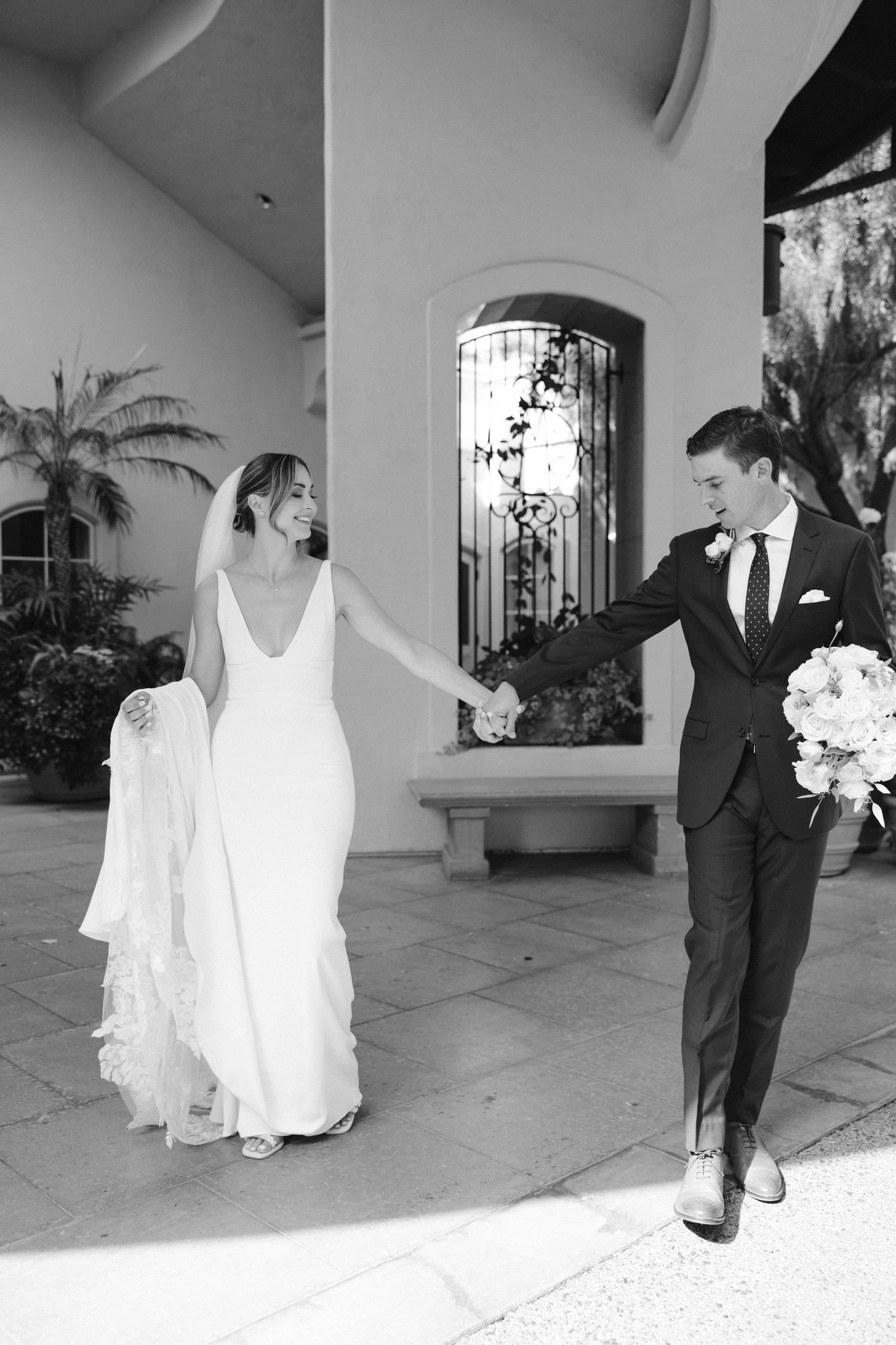 Black and white photo of bride and groom walking hand in hand on stone patio.