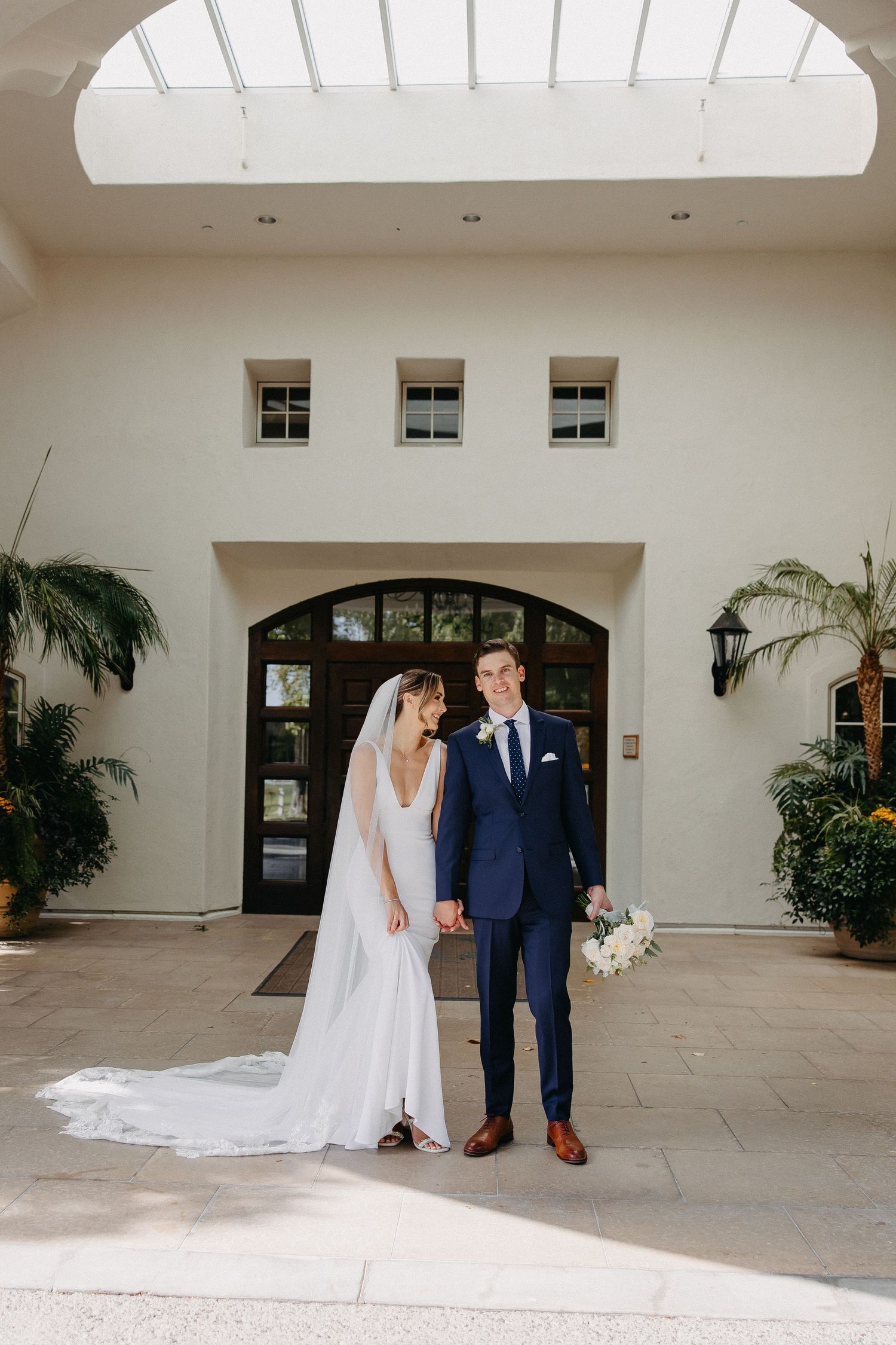 Wedding couple stands under skylight in Mediterranean-style building entrance.