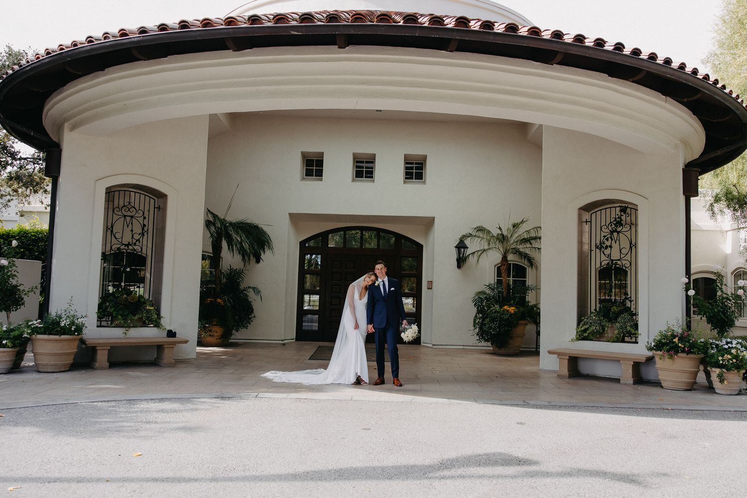 Wide shot of newlyweds under curved entrance of Mediterranean villa with palm trees.