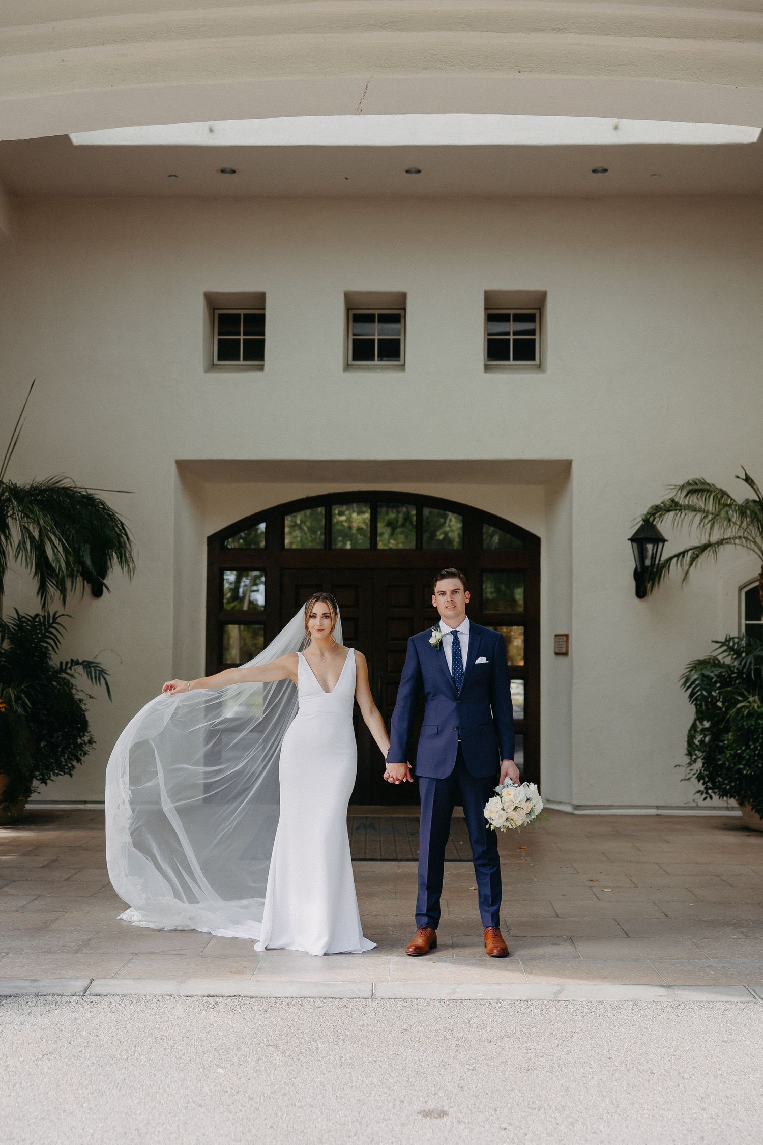 Bride's veil flows in breeze as couple poses under arched entryway with skylights.
