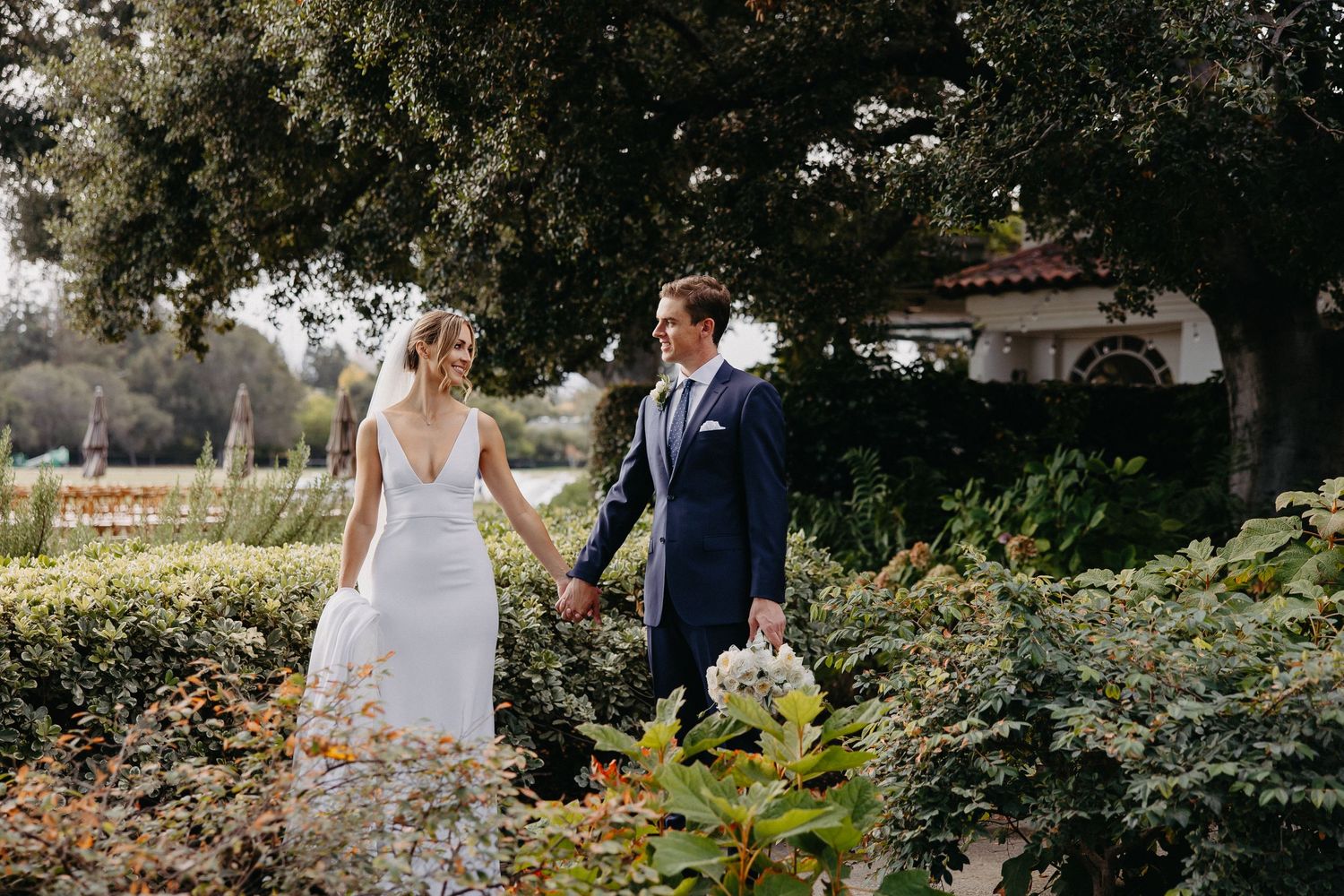 A romantic wedding couple walks hand in hand through a garden path surrounded by lush greenery.