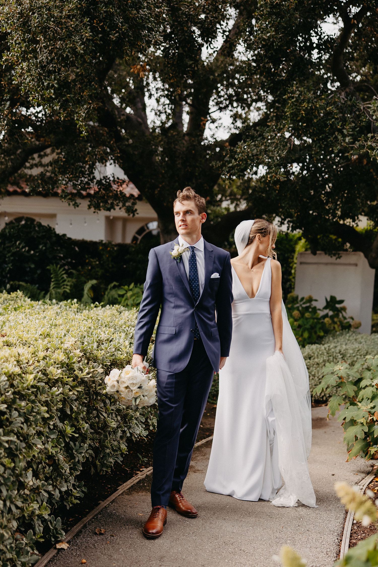 A newlywed couple strolls along a landscaped pathway with the bride in white and groom in navy.