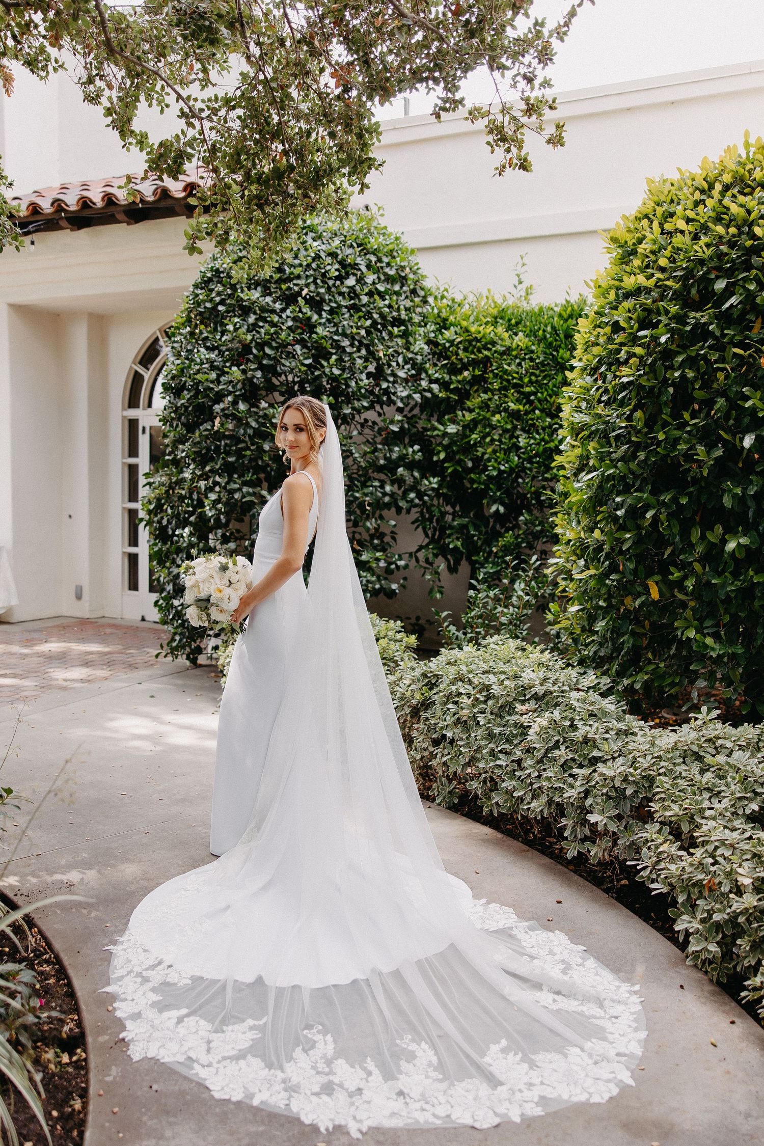 Wedding dress with dramatic cathedral train photographed against white architecture and garden backdrop.