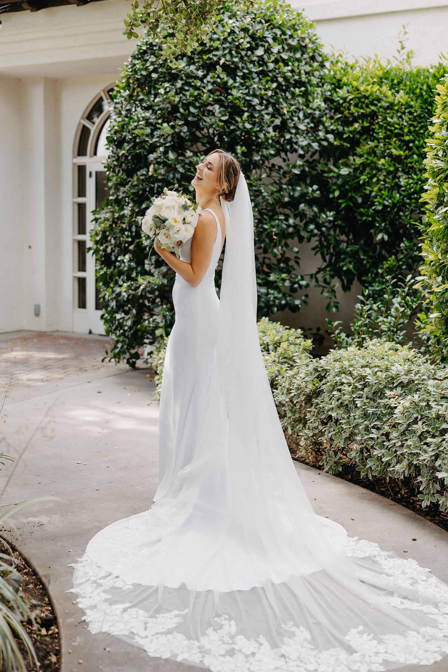 Bride in flowing white gown holds bouquet while standing in front of ivy-covered architecture.