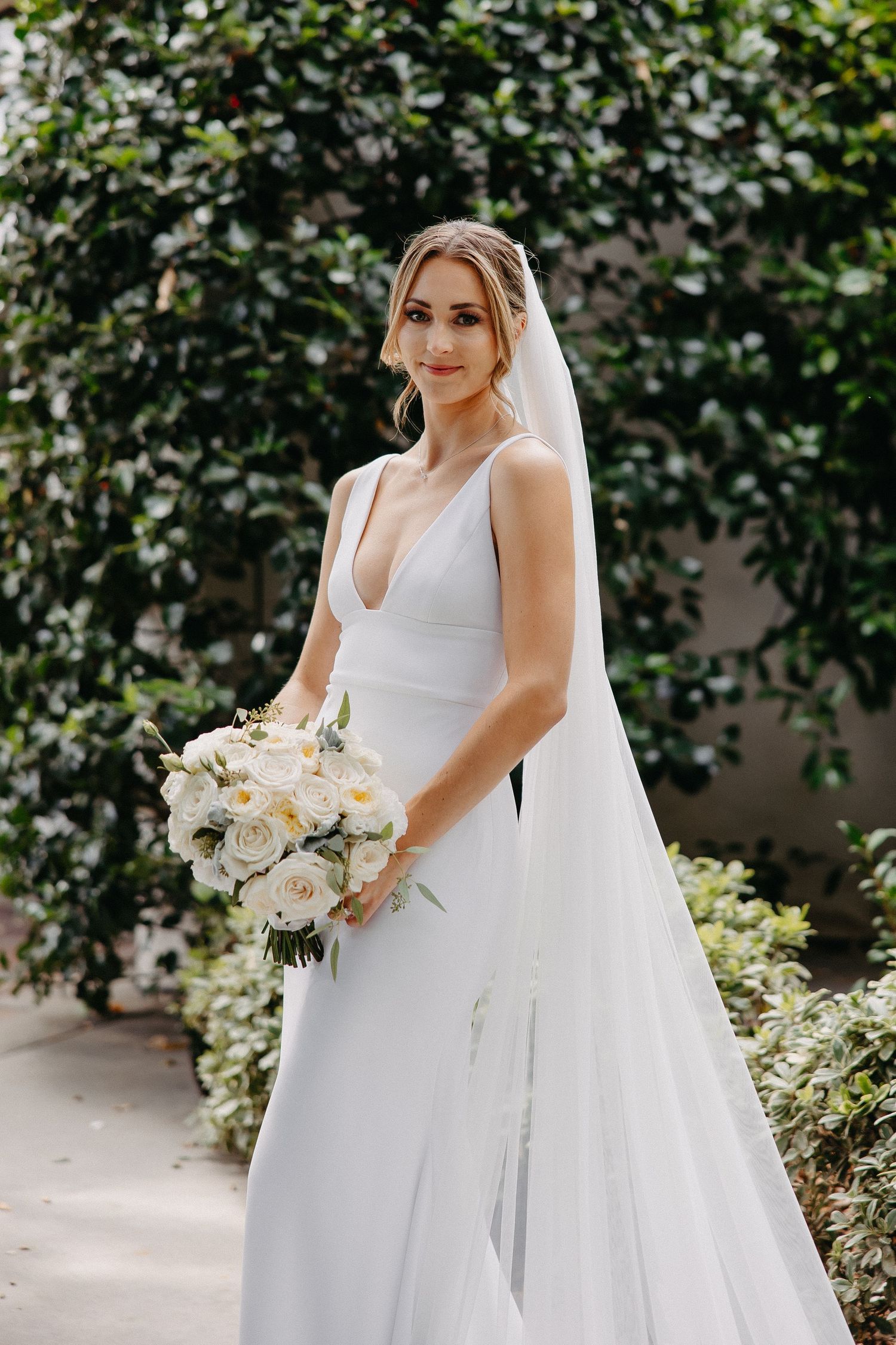 Elegant bride in sleek white gown holds bouquet of cream roses in garden setting.