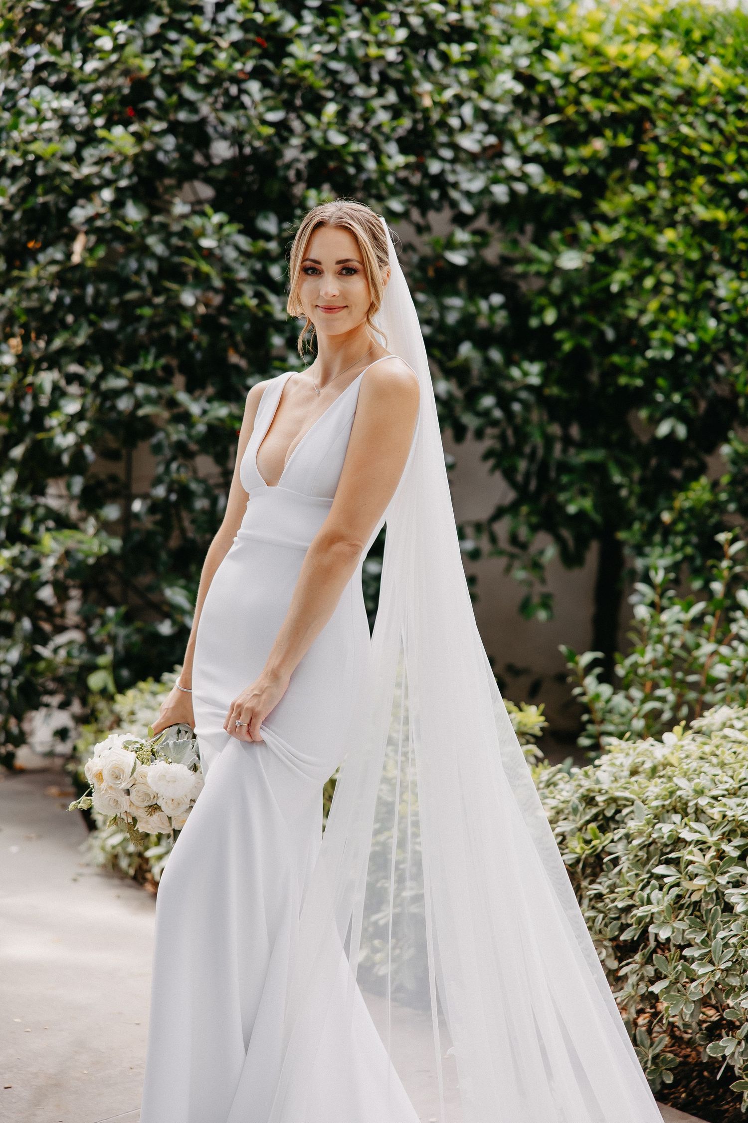 Elegant bride poses in fitted white gown with plunging neckline against garden greenery.