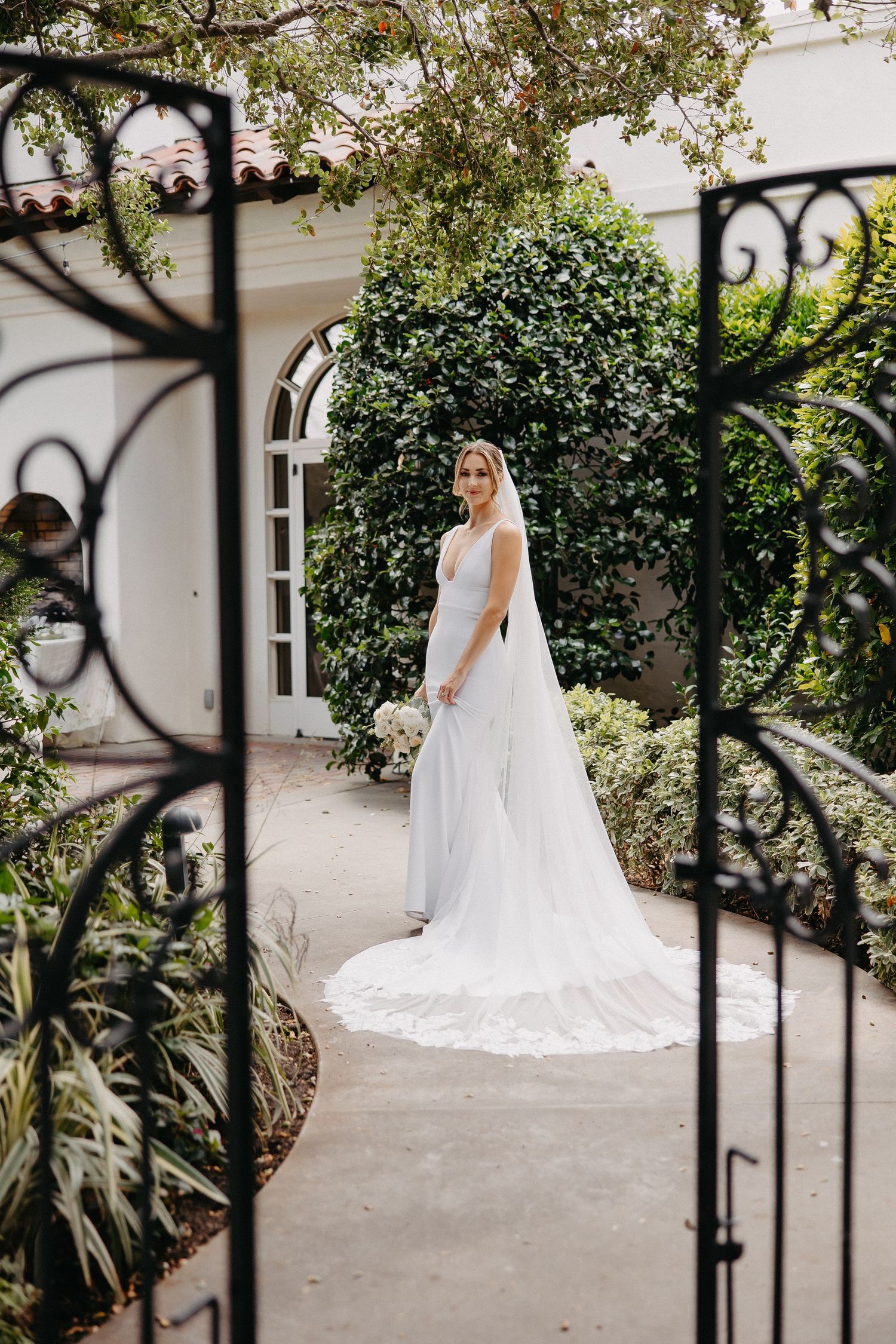 Elegant white wedding gown with flowing train viewed through decorative garden gate at outdoor venue.